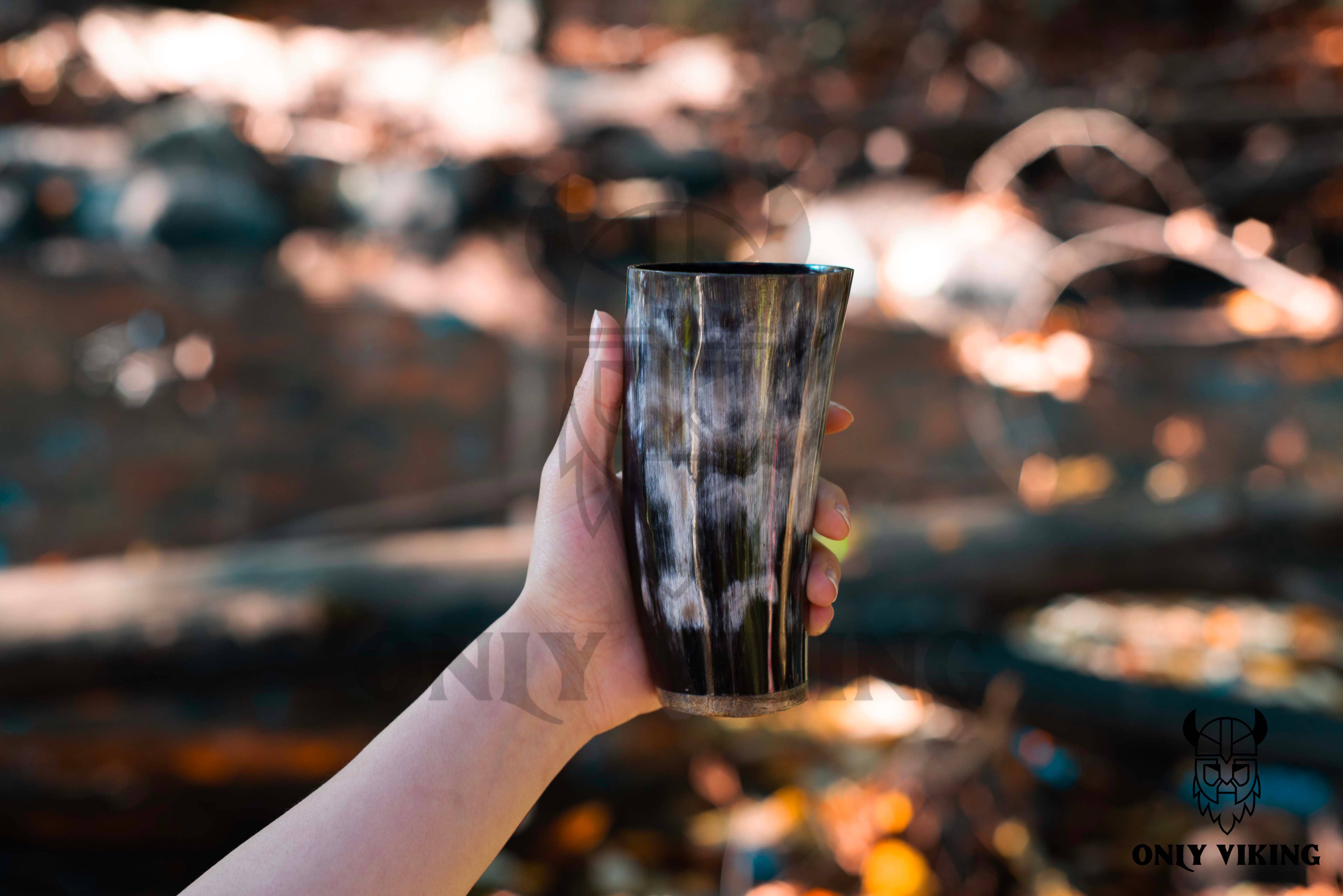 A hand holds the Drinking Horn: Beer Stein 6 Inch outdoors, with blurred trees and sunlight in the background.