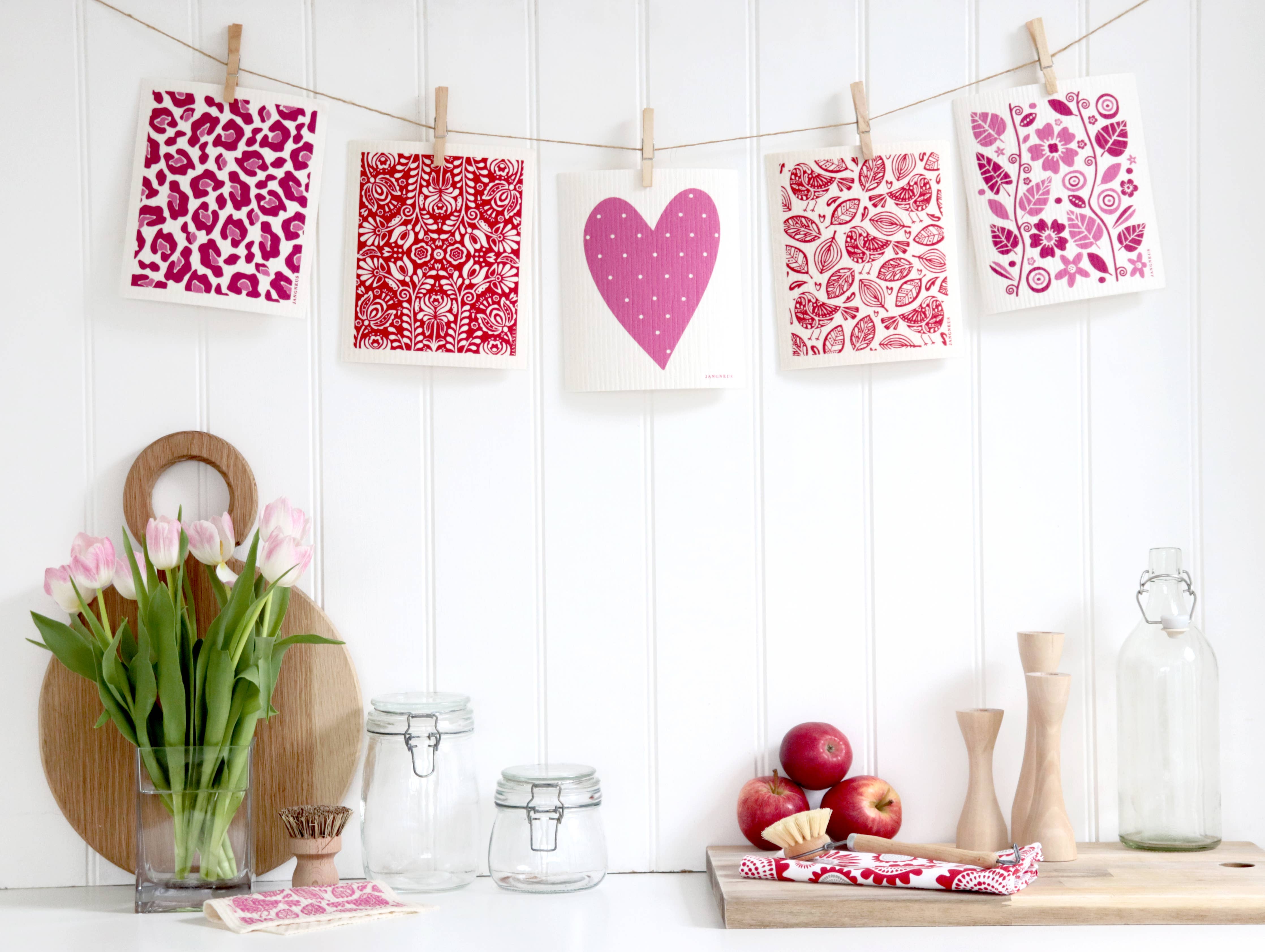 A string with red Swedish dish cloths featuring a large pink heart hangs by clothespins. Below are Scandinavian kitchen accessories like tulips, glass jars, a cutting board, apples, and wooden utensils—all set against a white background.