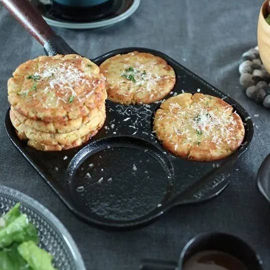 Three golden-brown pancakes with cheese and herbs cook in the Skeppshult Egg Pan With Walnut Handle 8 / 20 cm, while extra pancakes are stacked on a grey table nearby.