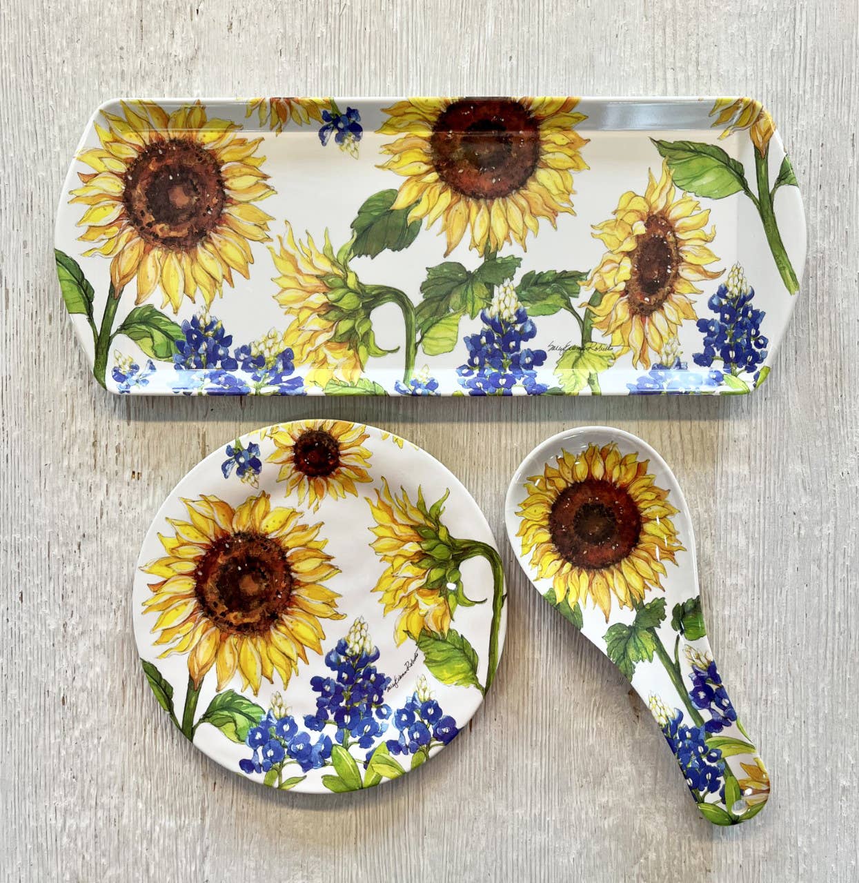 The Sunflower & Bluebonnets Floral Loaf Serving Charcuterie Tray, with matching round plate and spoon rest, features cheerful floral designs on bamboo—eco-friendly dinnerware ideal for gatherings, displayed on a light wooden surface.