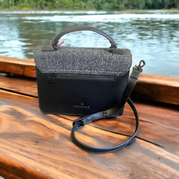 The Harris Tweed Large Satchel - Grey, featuring black leather and handwoven tweed with top handle and shoulder strap, rests on a wooden surface near the water, with trees in the background.