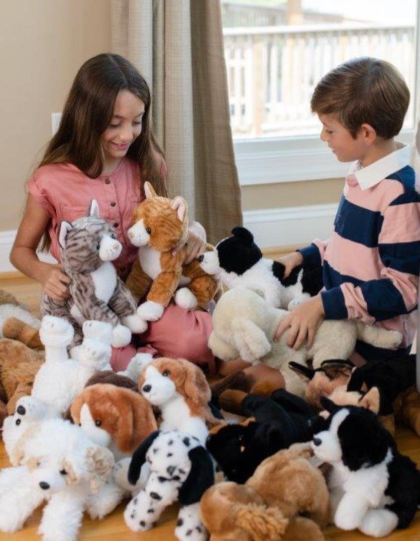 Two children sit on a wooden floor indoors, surrounded by various stuffed animal toys, including cats and a Plush German Shepherd Dog 12 tall stuffed animal.