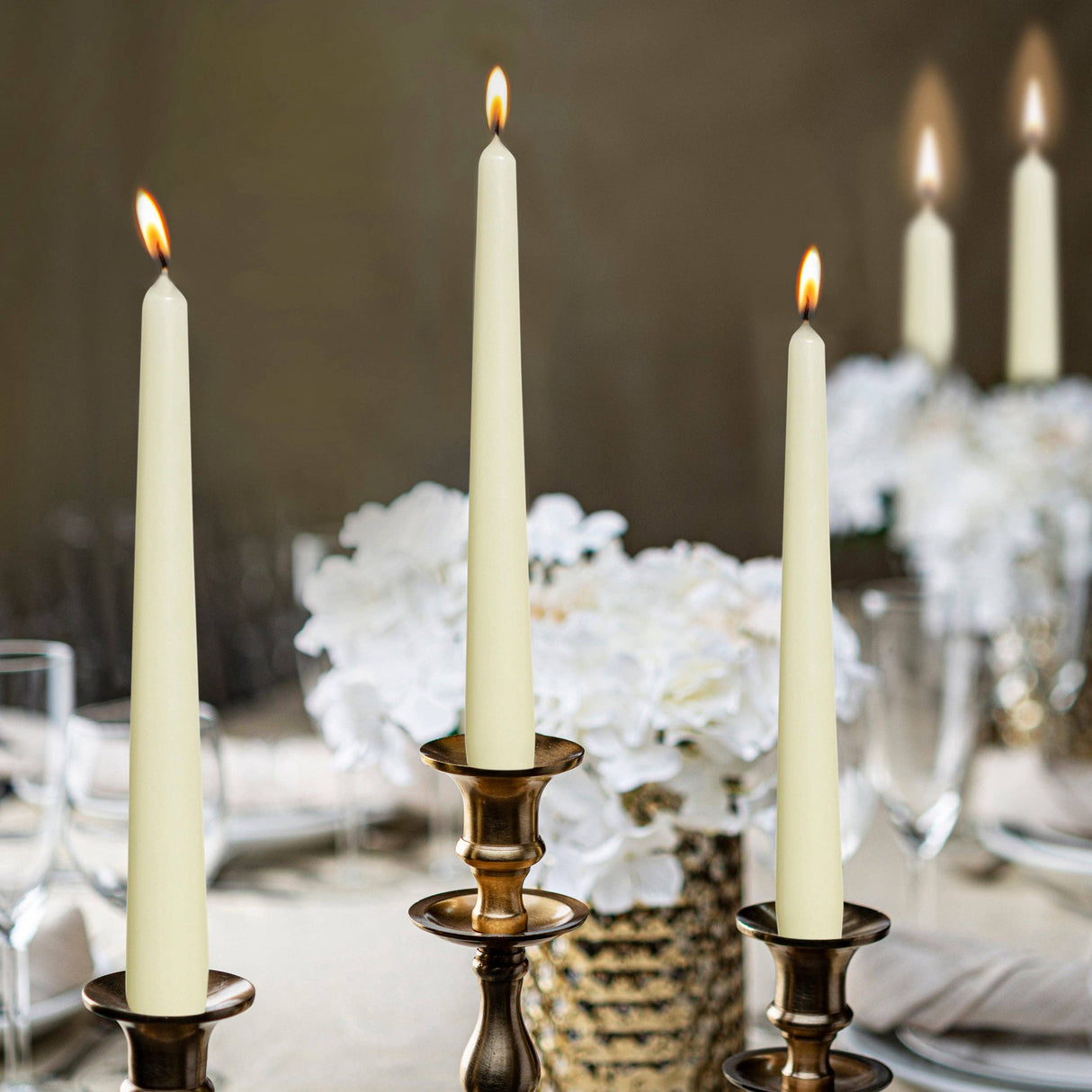 Three 10 inch unscented taper candles from the White, Red, or Ivory Tall Taper Candles 4 Pack sit in brass holders amid white flowers and glassware, creating an elegant and formal atmosphere with dripless charm.