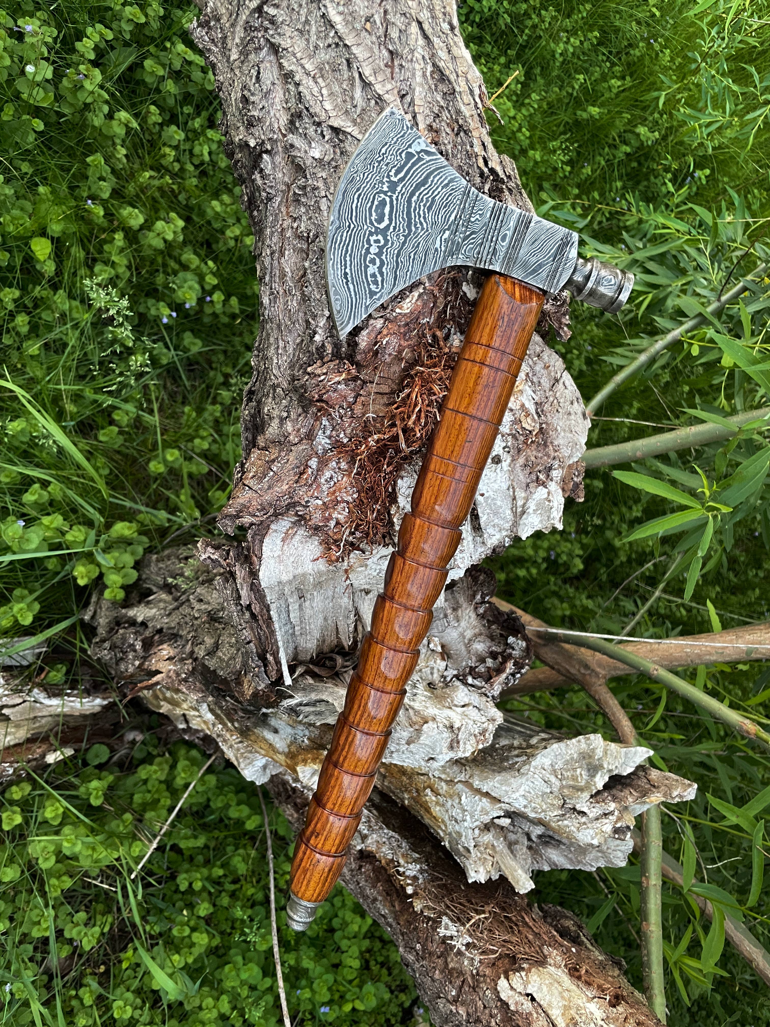 The Axe: Peace Pipe Axe Handmade with Damascus Steel features an engraved blade and carved wooden handle, resting on a fallen tree trunk amid lush greenery.