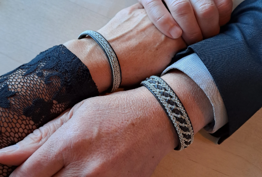Two people clasp hands on a table, each wearing the Lapland Sami Tennis Bracelet #2 in Cognac (19-20 cm); one is in black lace, the other wears a dark suit.