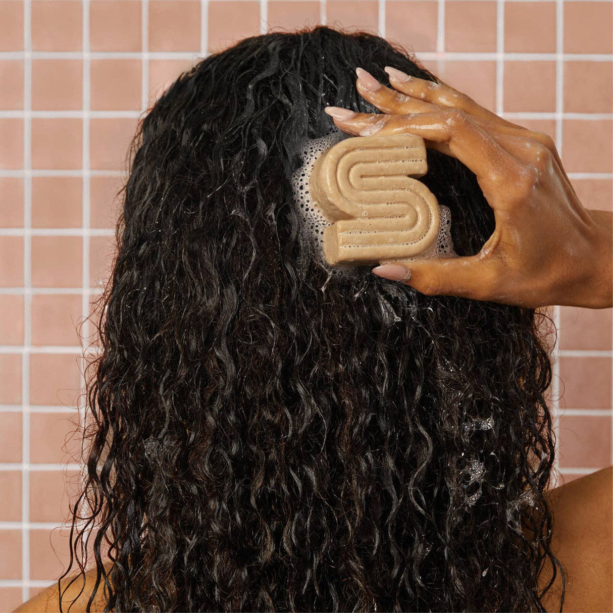A person with wet, curly hair washes their scalp using Kitsch’s Coconut Oil Shampoo & Conditioner Bar Combo for dry, damaged hair (2 x 2.7oz), enjoying hydrating care against a pink tiled wall.