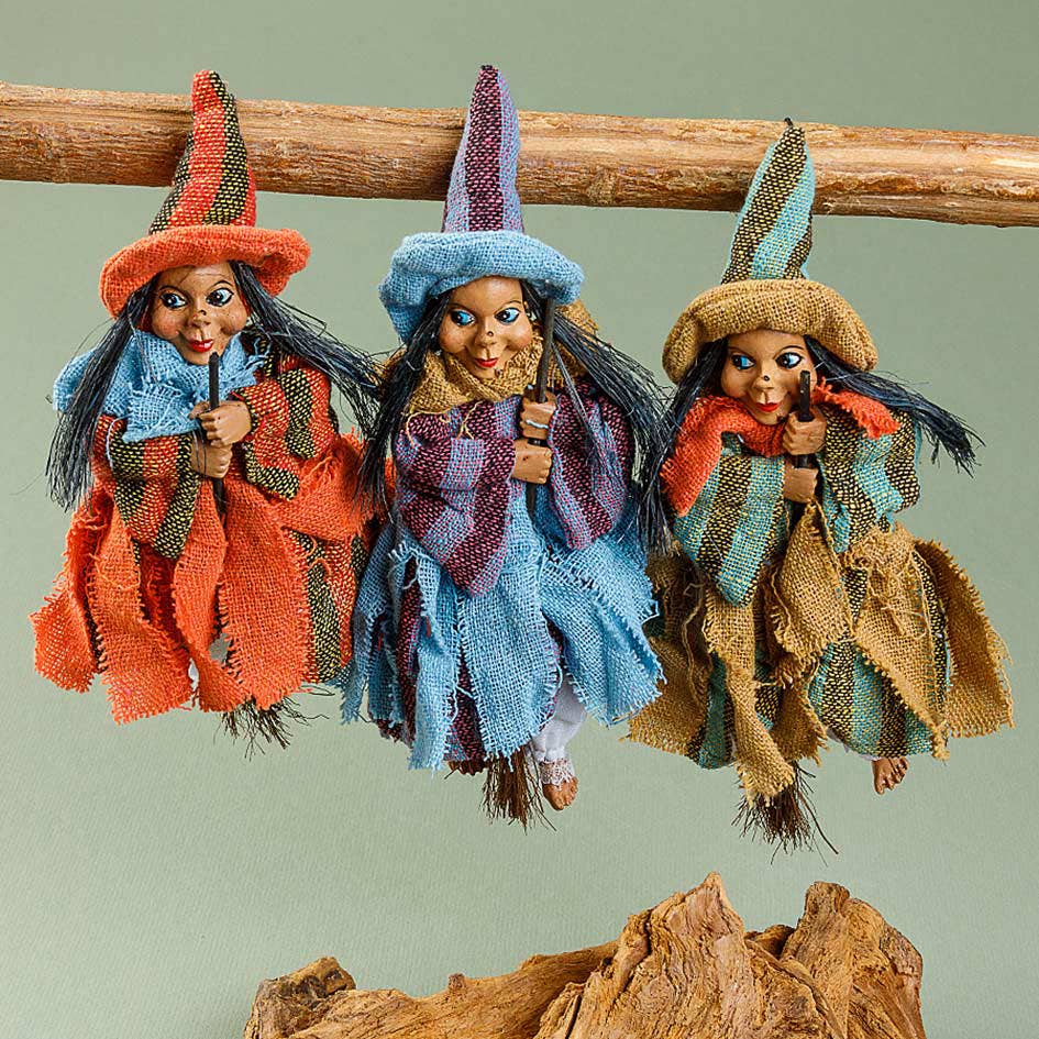 The Philippa Blue Kitchen Witch Doll features a colorful hat and cloak, long black hair, and an expressive face. Handmade in Germany, this good luck charm sits on a wooden branch against a plain background.