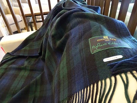 A green and navy tartan wool scarf with fringe edges, reminiscent of the Blanket: Black Watch Highland Blanket, is draped over a surface. A label reading Patrick King Highlands Collection is visible.