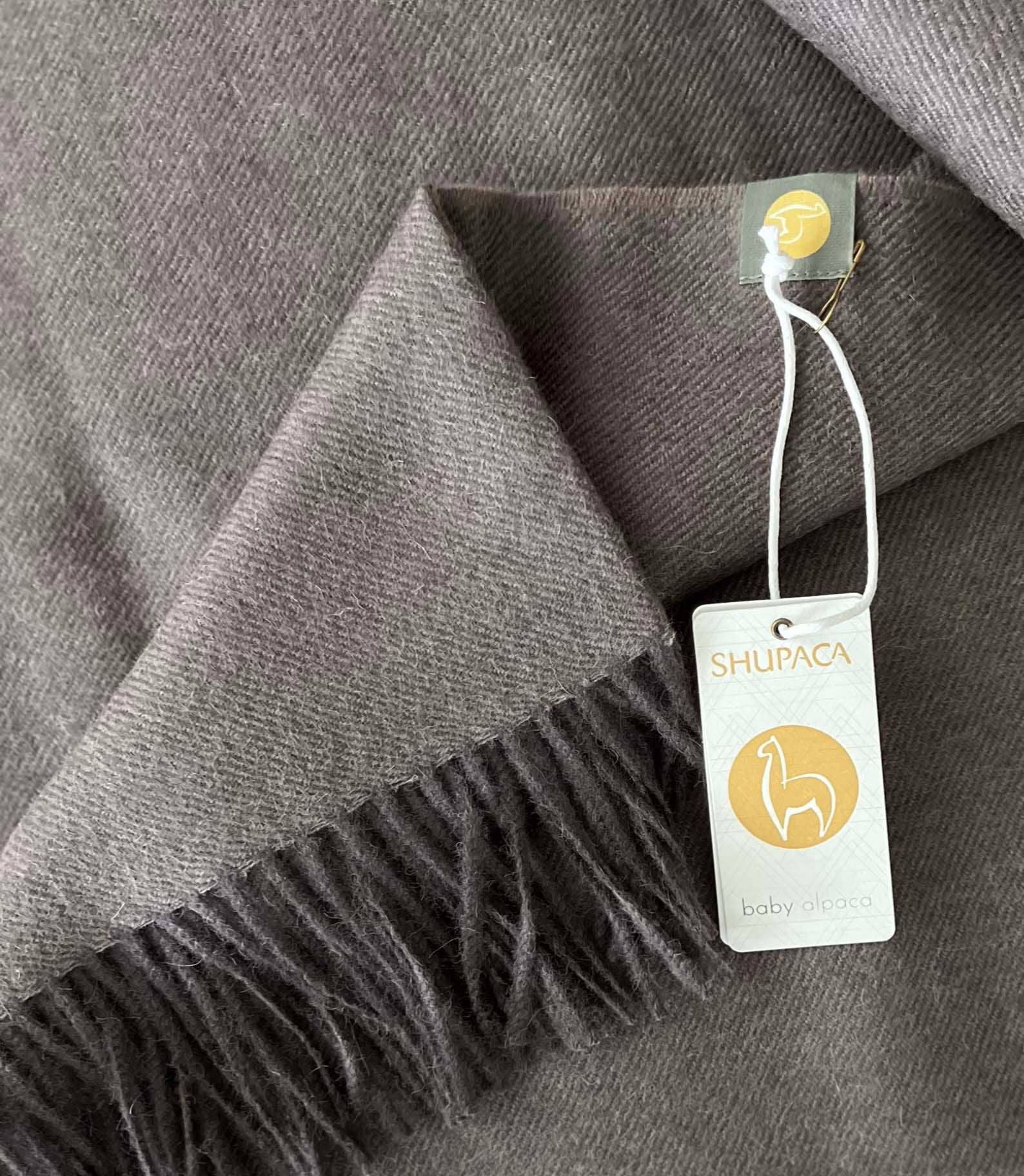 The "Scarf: 100% Baby Alpaca Scarf - Truffle" provides luxurious warmth with its finest fiber, featuring fringe and a Shupaca label, and includes a tag that reads baby alpaca.