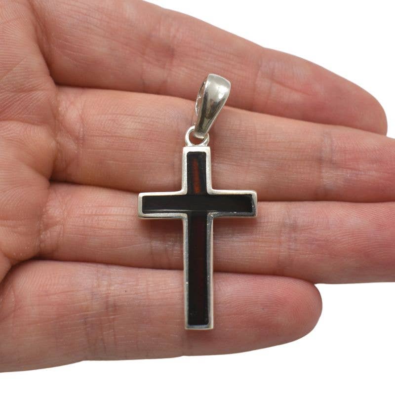 A hand holding the Cherry Amber Sterling Silver Cross Pendant, a black cross with a silver border, crafted from elegant sterling silver.