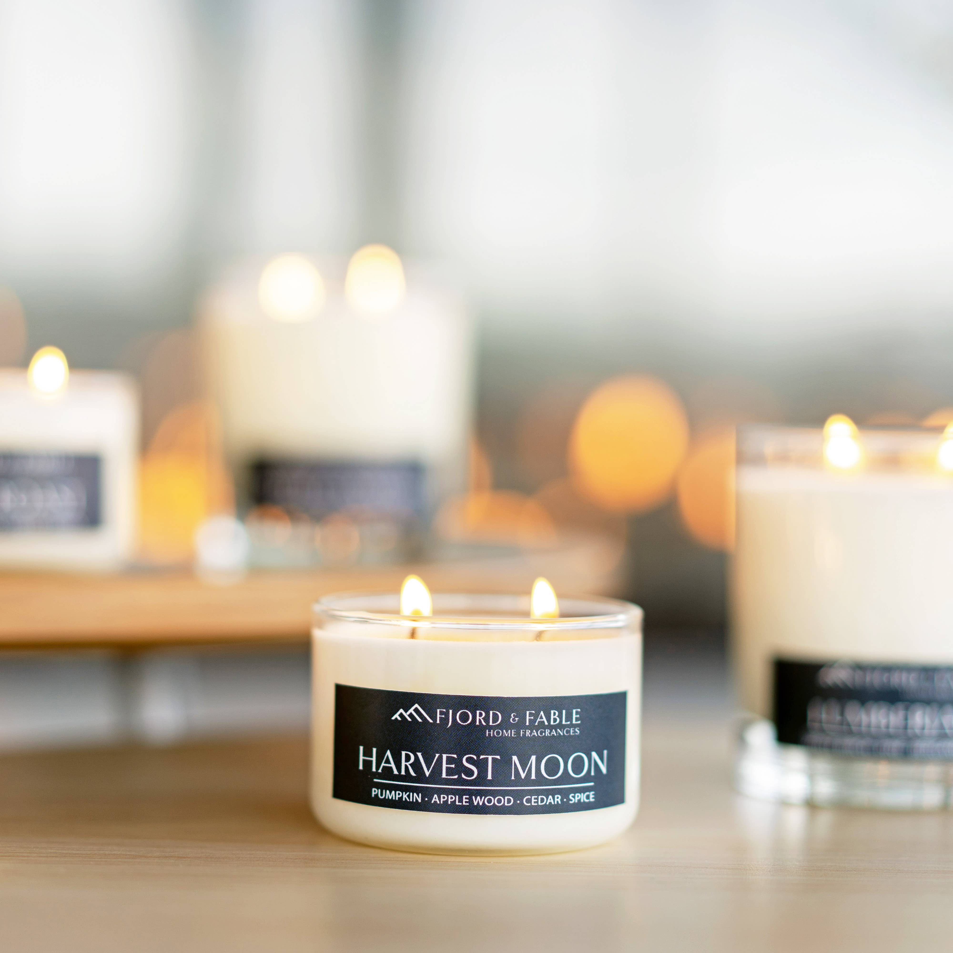 A lit Cedar Tobakk hand-poured soy candle sits on a table with other candles in the background; its label features notes of tobacco, amber, patchouli, and peppercorn for a rich, aromatic fragrance.