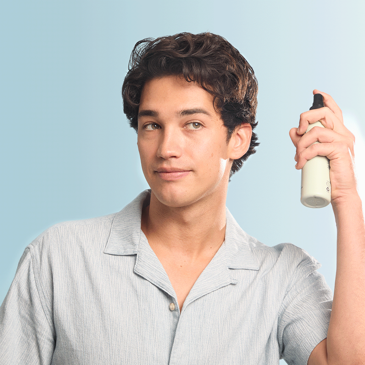 A young man with wavy dark hair, dressed in a light blue shirt, confidently holds a bottle of KITSCH Spray: Texturizing Sea Salt Hair Spray with Rosemary & Biotin (6 fl oz) near his head, gazing to the side against a soft blue background.