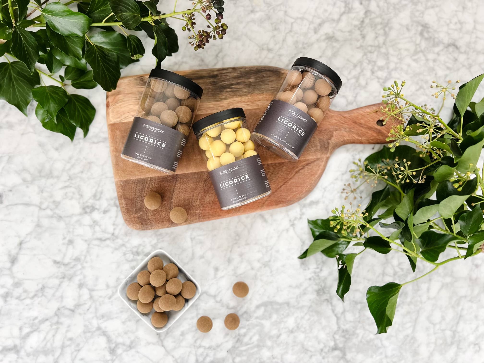 Three jars of Schottinger Licorice with White Chocolate & Passionfruit (250g) sit on a wooden cutting board, with loose candies nearby and green leafy plants surrounding them on a marble surface.
