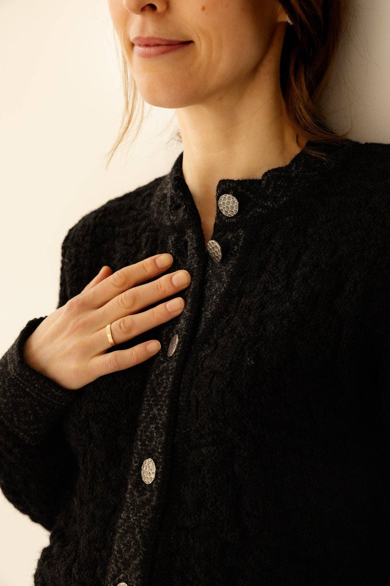 A person wearing the Bryggen Women's Norwegian Cardigan in white with silver buttons rests their gold ring-adorned hand on their chest. Only the lower face and upper torso are visible.