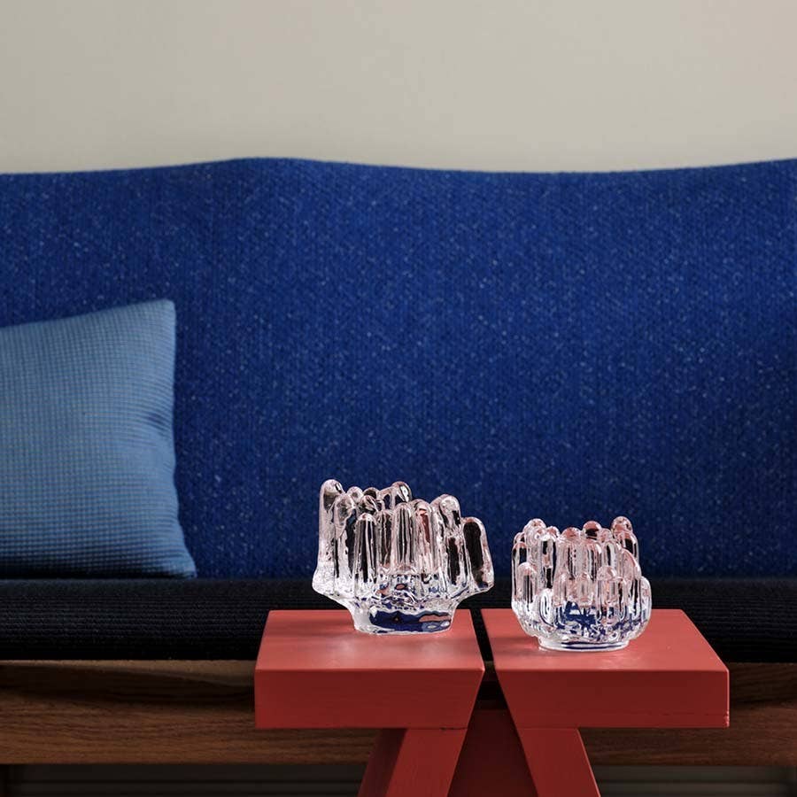 A KostaBoda Polar Votive Clear Small (H-86mm x D-95mm) with jagged edges sits on a red table in front of a blue upholstered sofa with a blue pillow.
