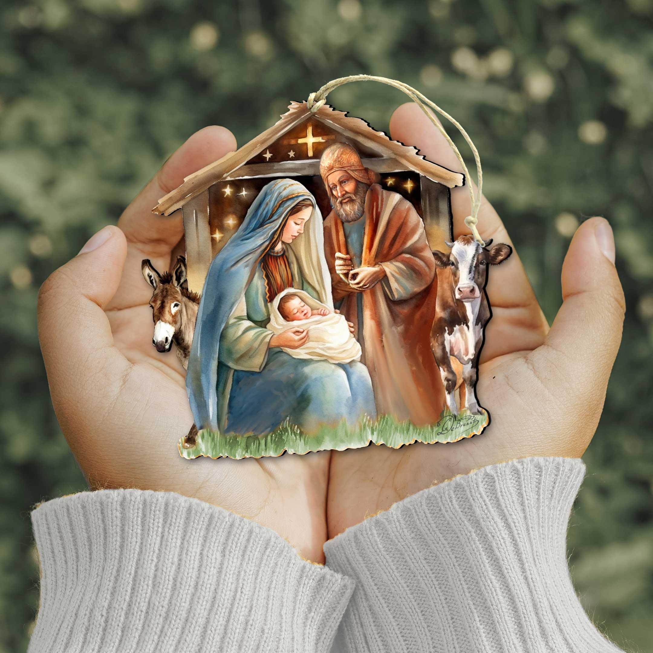 A person holds the "Born Under Bright Star Nativity" wooden ornament by G. DeBrekht (approx. 5 x 4.75 in), featuring Mary, Joseph, baby Jesus, a cow, and a donkey, with a blurred green background.
