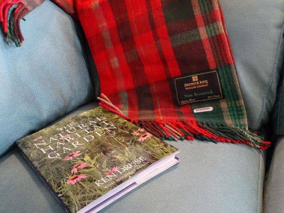 A book titled The Natural Habitat Garden rests on a blue sofa beside a New Brunswick Deluxe Highland Tartan Blanket, 100% wool, measuring 69" x 62", with its label visible.