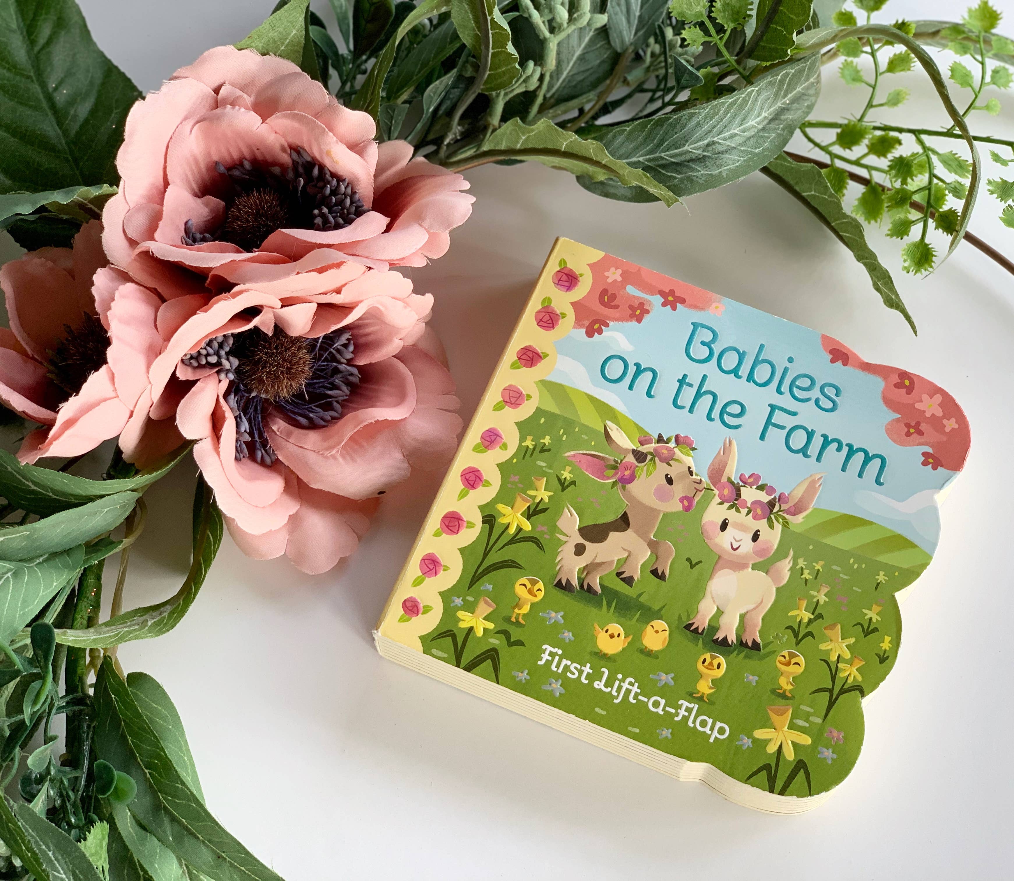 The Book: Babies on the Farm Lift-a-Flap Board Book rests on a white surface, surrounded by pink flowers and green foliage.