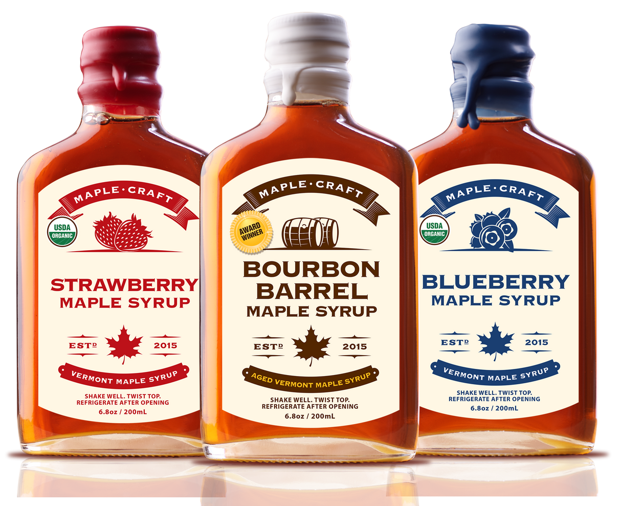 Three bottles of organic, refined sugar-free maple syrup stand side by side with labels: Strawberry - Summer Favorites Maple Syrup (6.8 oz), Bourbon Barrel, and Blueberry. Each features a maple leaf insignia and the year 2015.