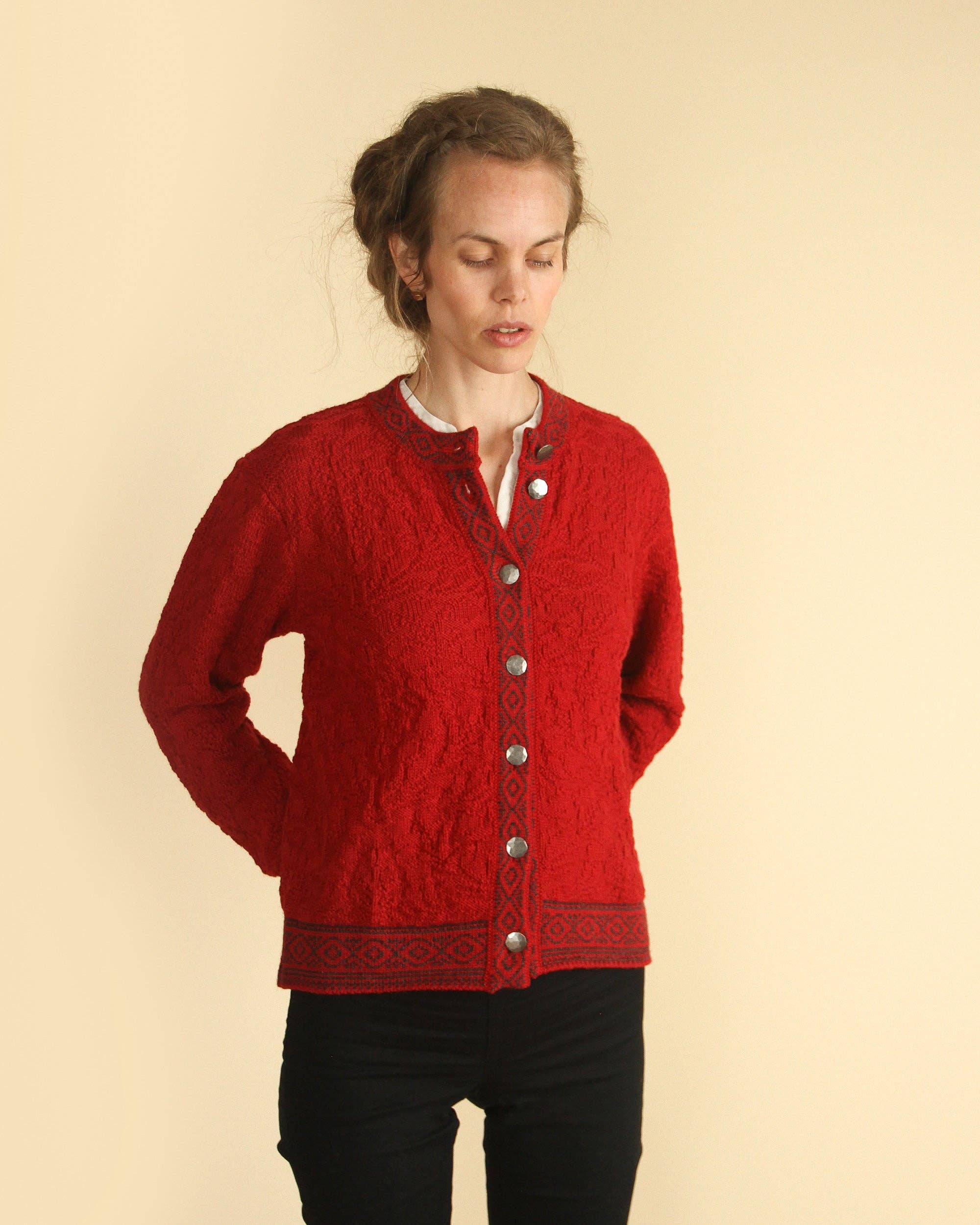 A woman with light brown hair wears the Bryggen Women's Norwegian Cardigan in white, featuring black trim, and stands against a plain beige background with her hands behind her back.