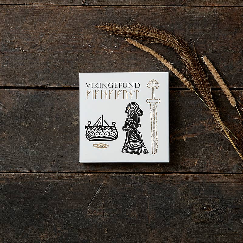 A white box labeled Card Pack: Viking Finds by Koustrup & Co is adorned with Viking motifs like a ship, a woman, and a sword. It rests on a wooden surface, surrounded by dried grass.