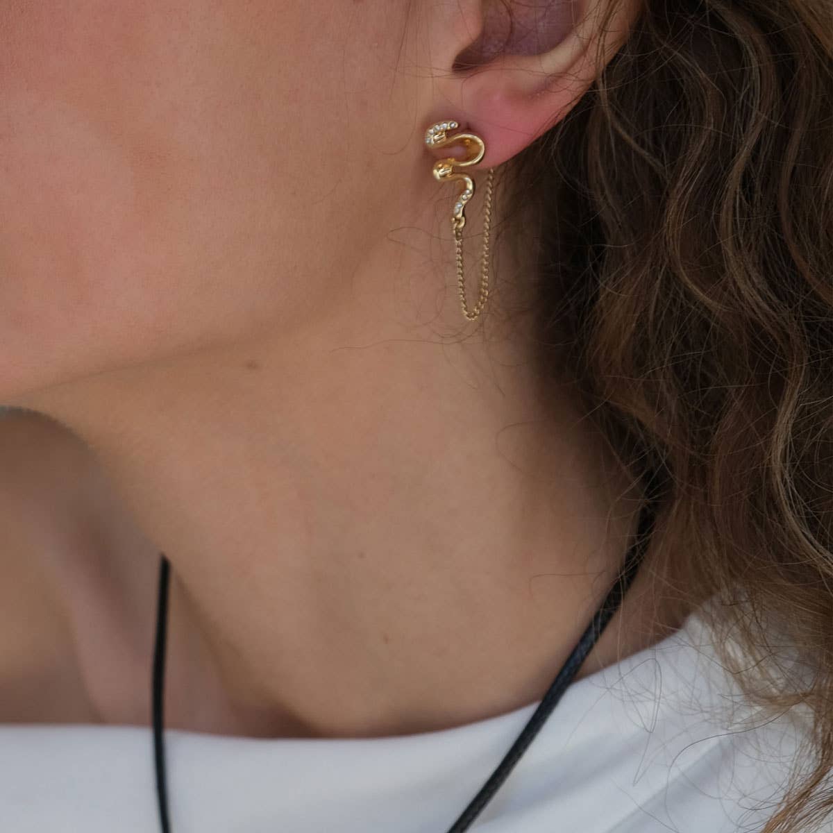 Close-up of a person with wavy brown hair in a white top wearing A&C Oslo Gold Stud Earrings with Chain, part of our blowing ribbon collection. These snake-shaped earrings are made in Norway.