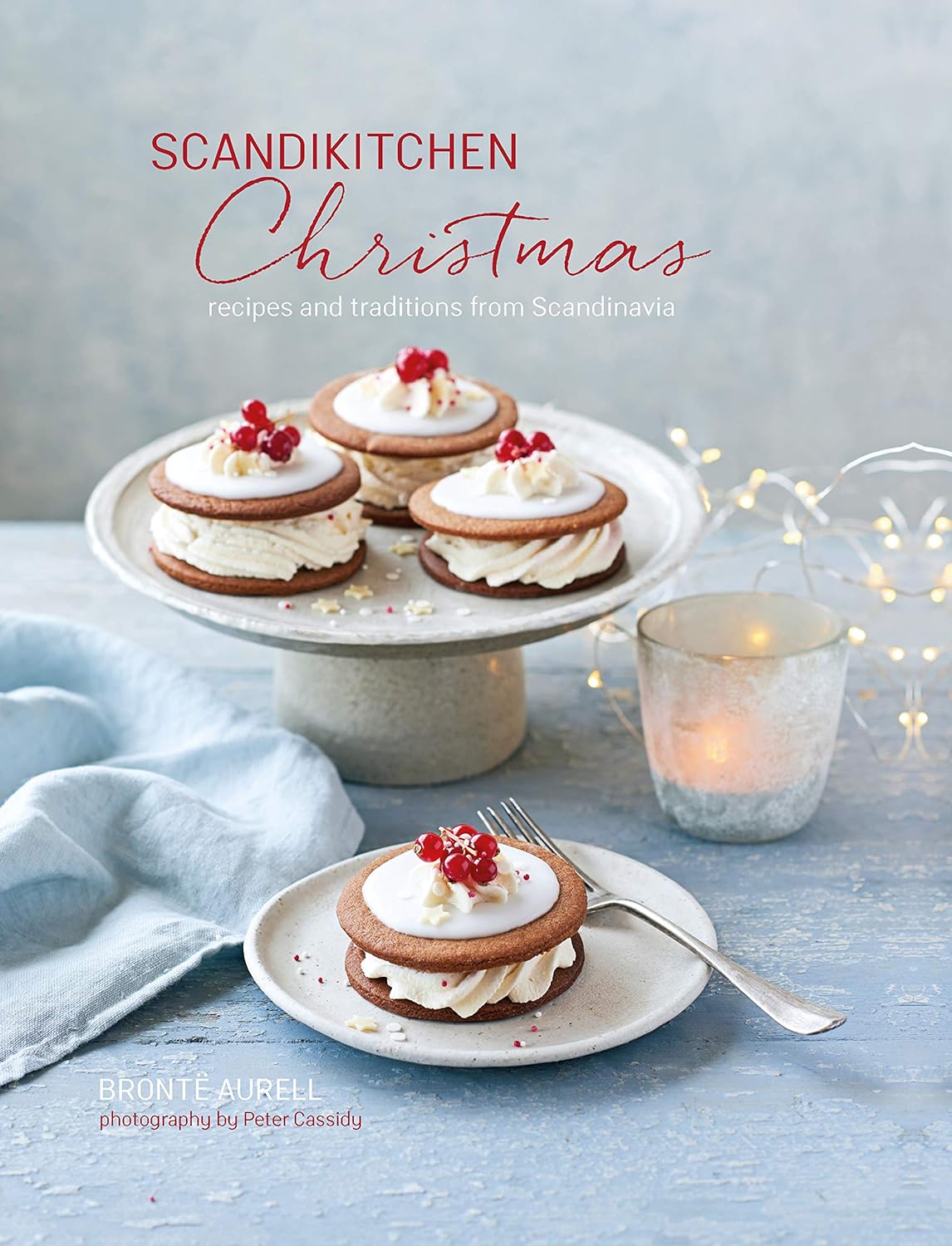 A book called "ScandiKitchen Christmas: Recipes and Traditions from Scandinavia" features a plate of festive cookies adorned with cream and red berries, capturing the cozy essence of Scandinavian Christmas traditions, all illuminated by a glowing candle.