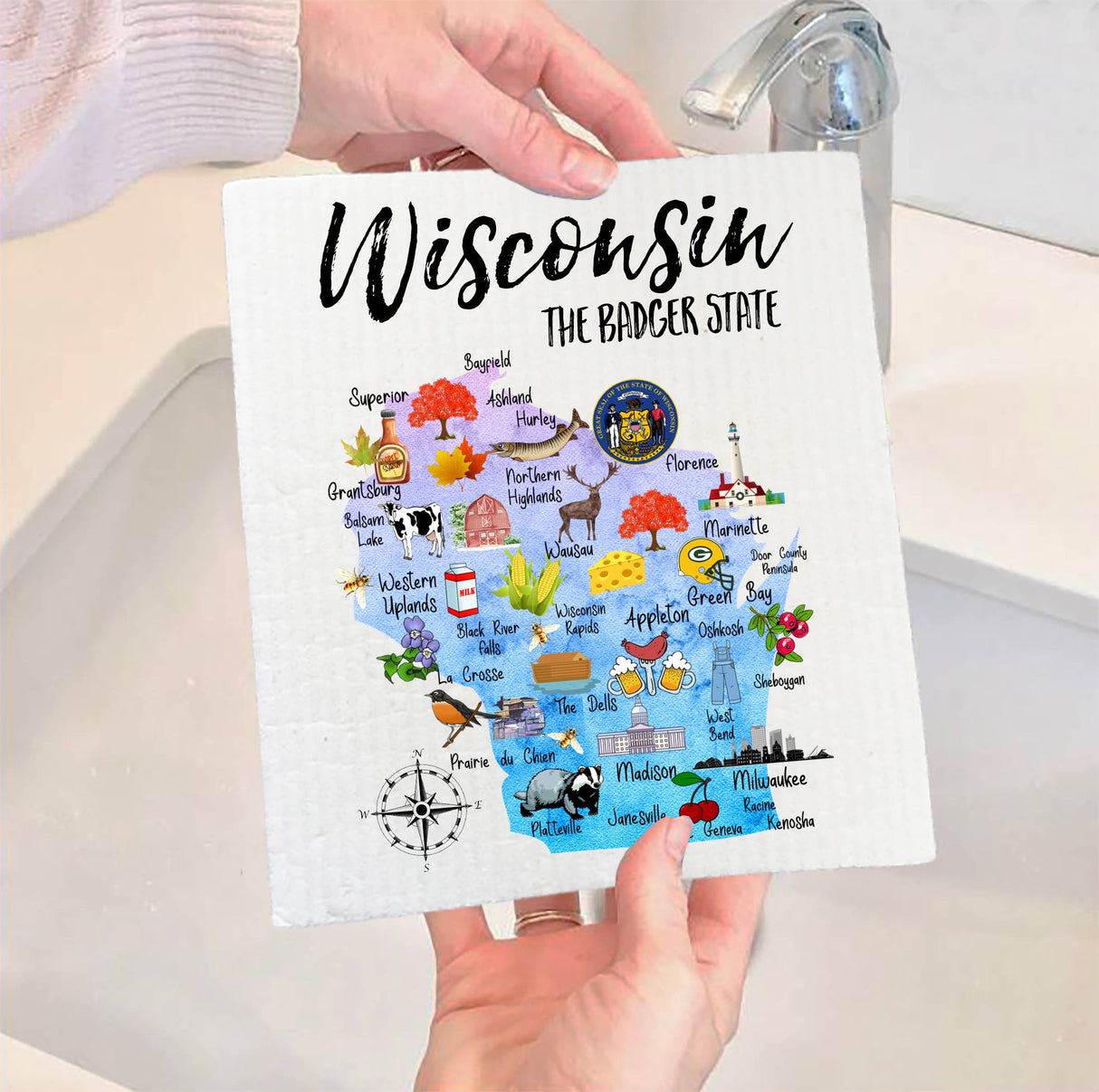A person holding a Swedish dish cloth featuring a colorful illustrated map of Wisconsin, labeled as The Badger State and showcasing various cities and state symbols.