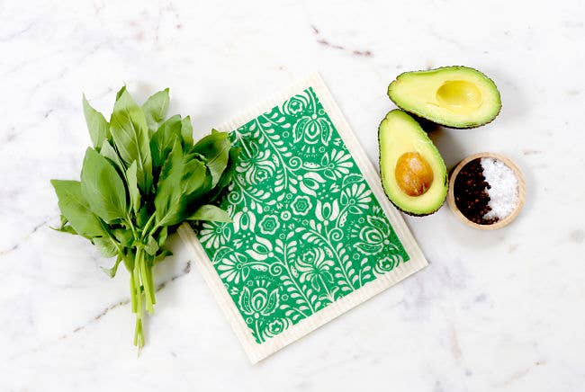 A collection of fresh greens, a sliced avocado, and a small bowl of salt and pepper sit on the Scandi Bloom - Green dish cloth. This biodegradable and absorbent cloth beautifully complements the marble surface, enhancing both style and sustainability in your kitchen setup.