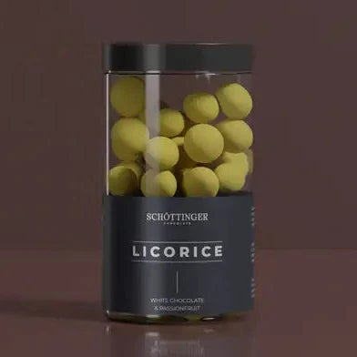 A clear jar labeled “Schottinger - Licorice with White Chocolate & Passionfruit (250g)” holds yellow-coated licorice candies, featuring a dark lid and label that highlights their unique white chocolate and passionfruit flavor.