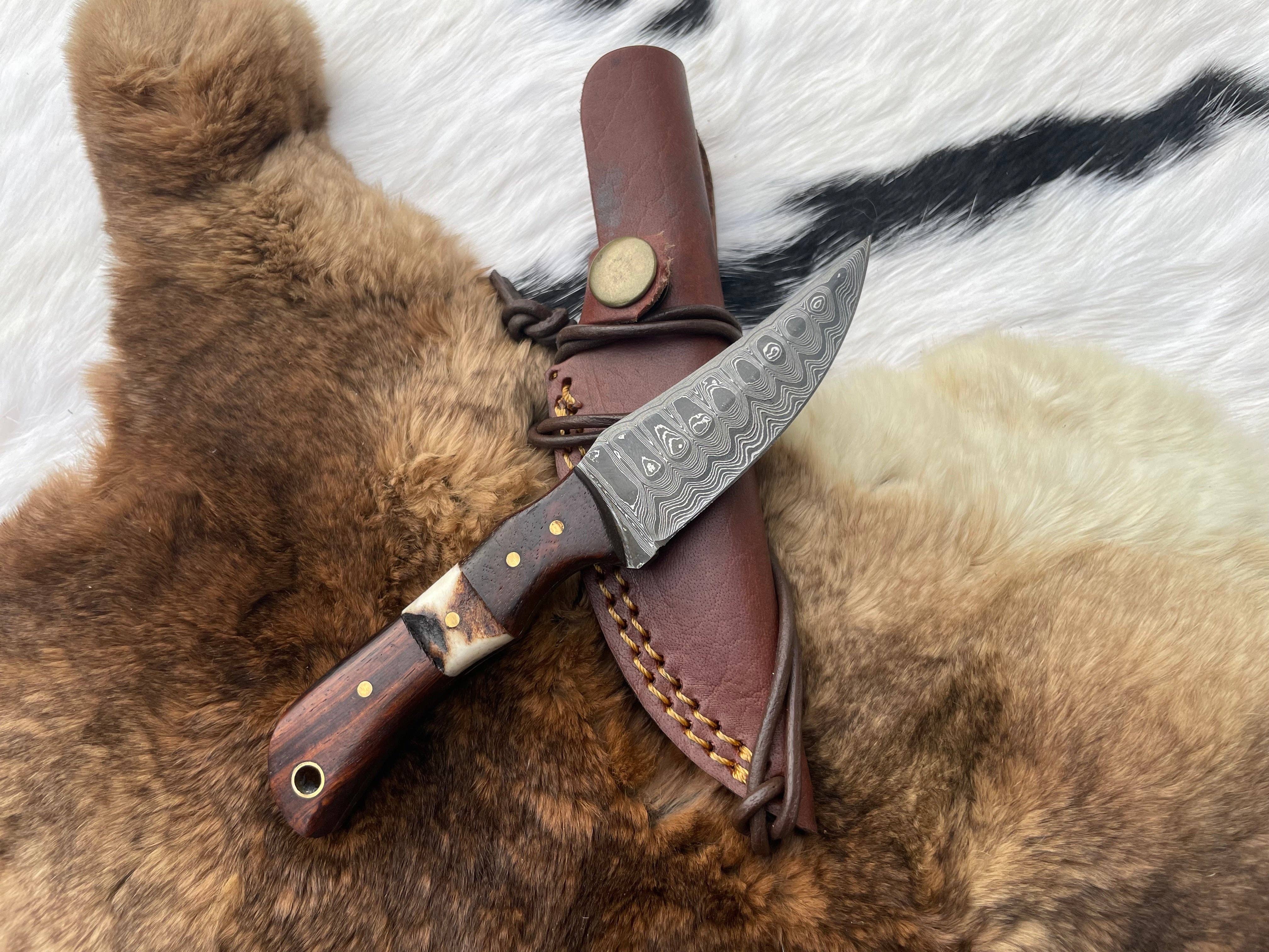 The Damascus Steel Skinner Knife with Rosewood & Stag Grip rests on a leather sheath atop animal fur, its patterned blade gleaming elegantly.