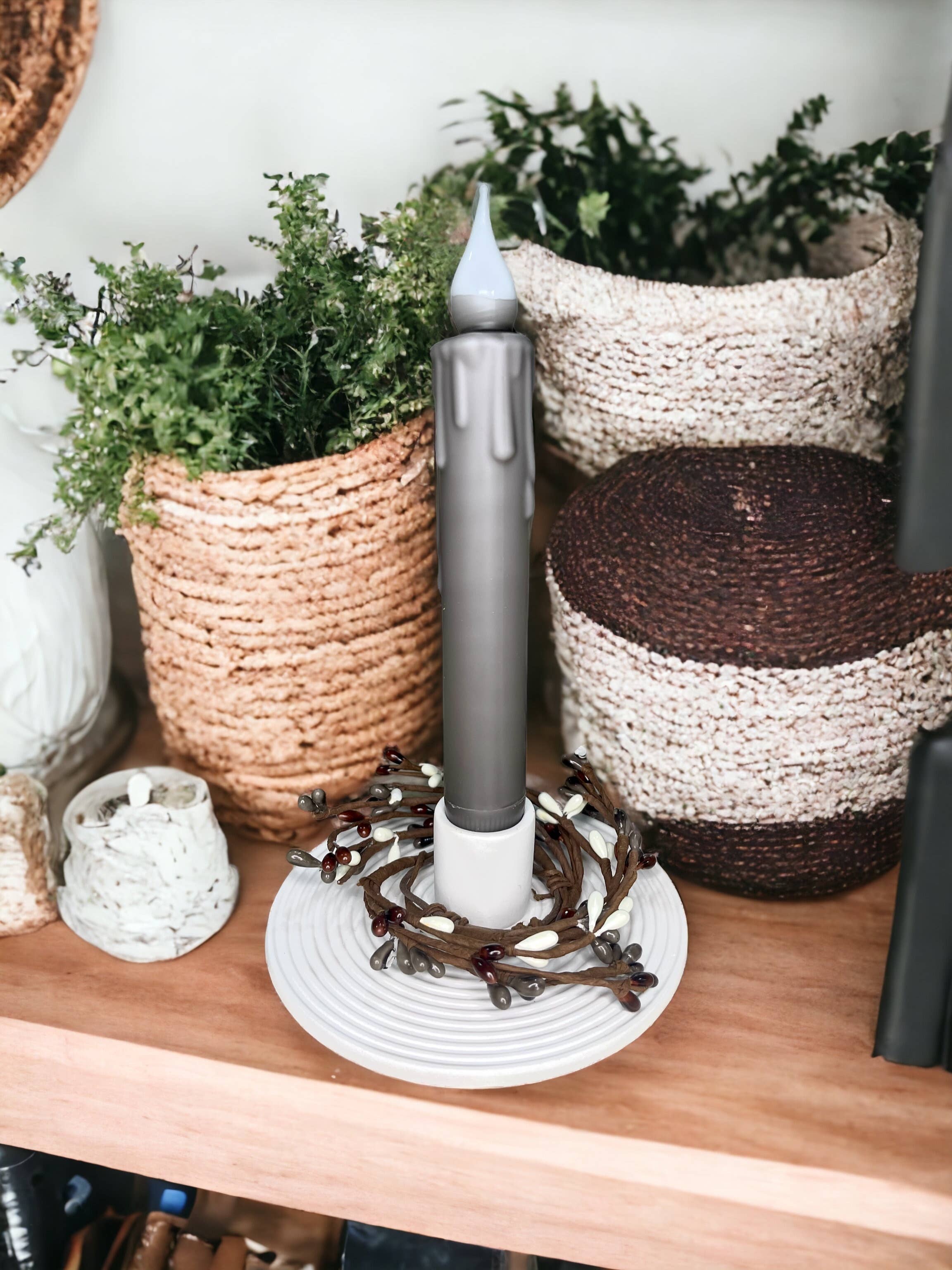 A tall, dark candle housed in the Modern White Porcelain Taper Holder sits amidst decorative vines with berries, forming an elegant tabletop decoration on a wooden shelf adorned with woven baskets and greenery. Ideal for adding charm to wedding events.