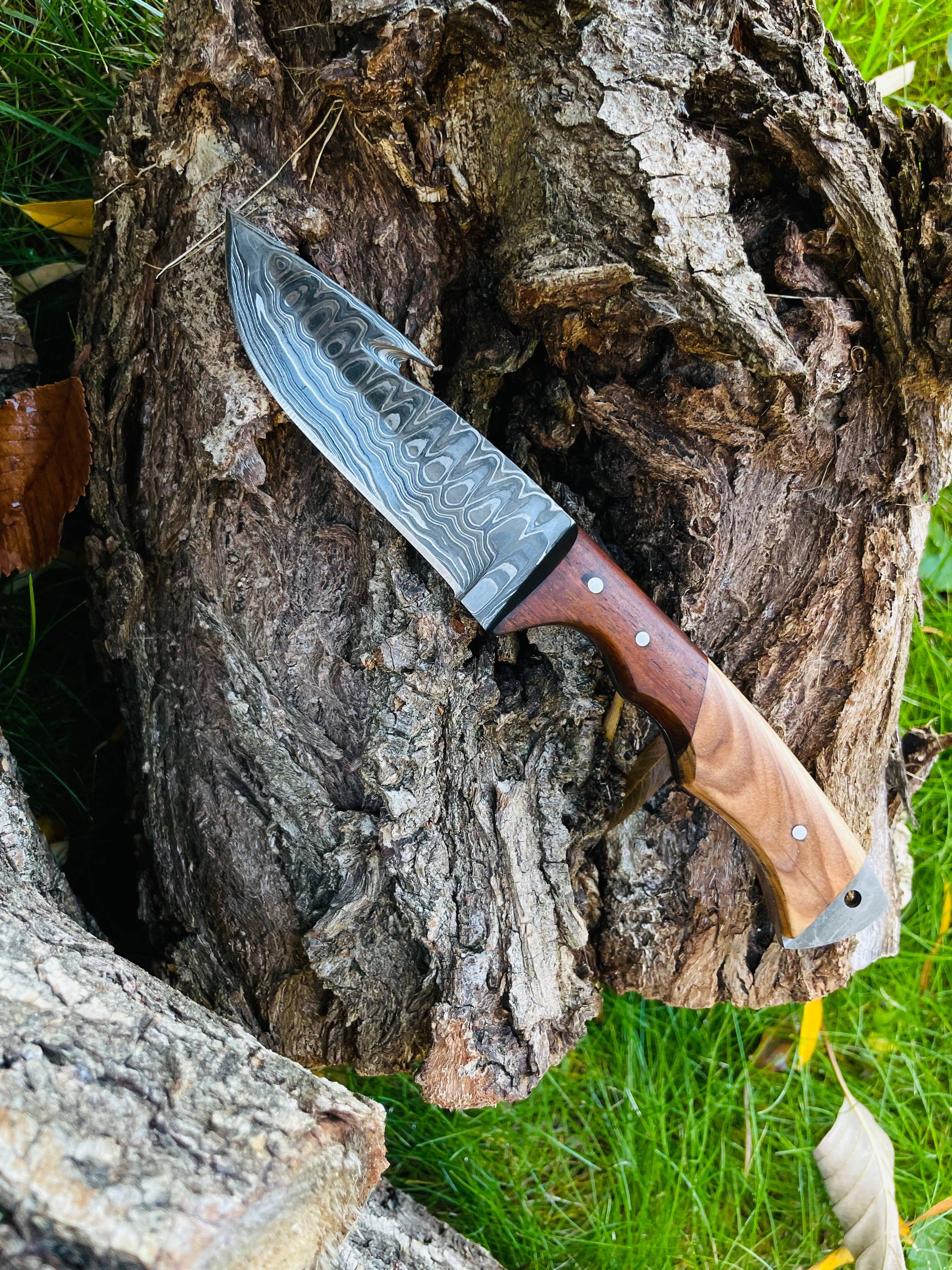 The Knife: Damascus Steel Guthook Skinning Knife with a patterned blade and wooden handle rests on rough tree bark outdoors, with green grass in the background.