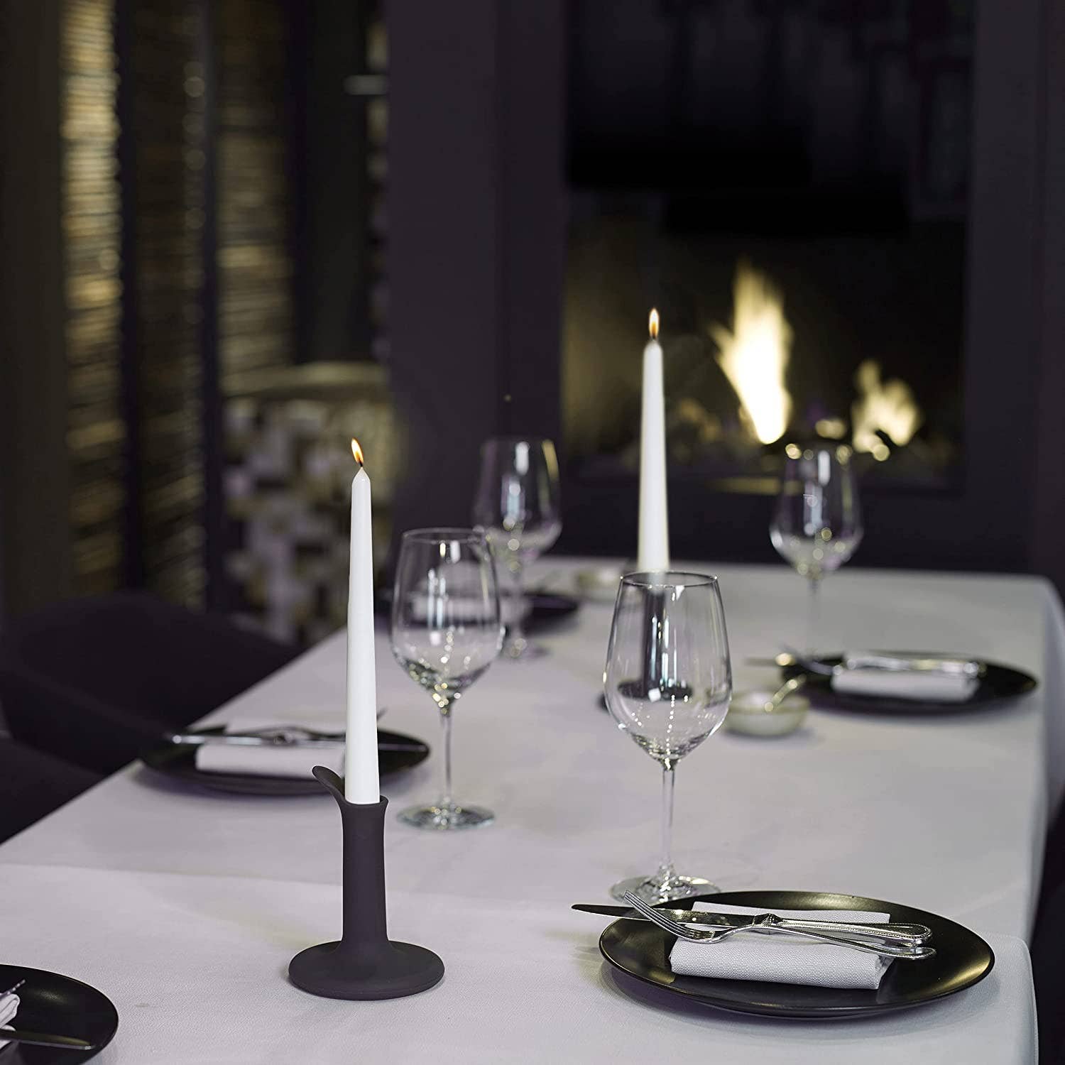 A dining table set with black plates, silverware, wine glasses, and two lit white 10 inch taper candles from the Unscented Taper Candles 4 Pack, with a fireplace burning in the background.
