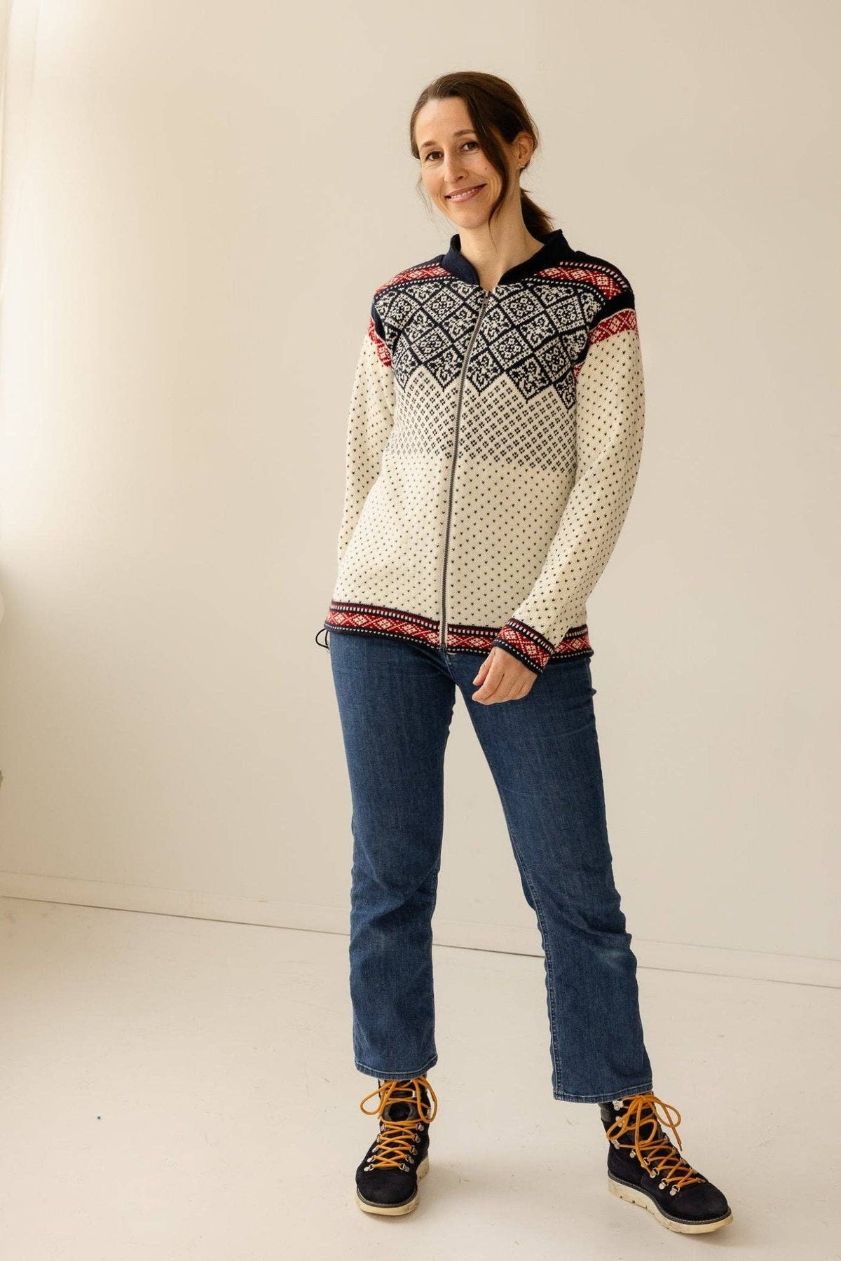 Woman wearing a white, fleece-lined Norwegian wool cardigan with a Kvam design yoke, blue jeans, and dark hiking boots with orange laces.