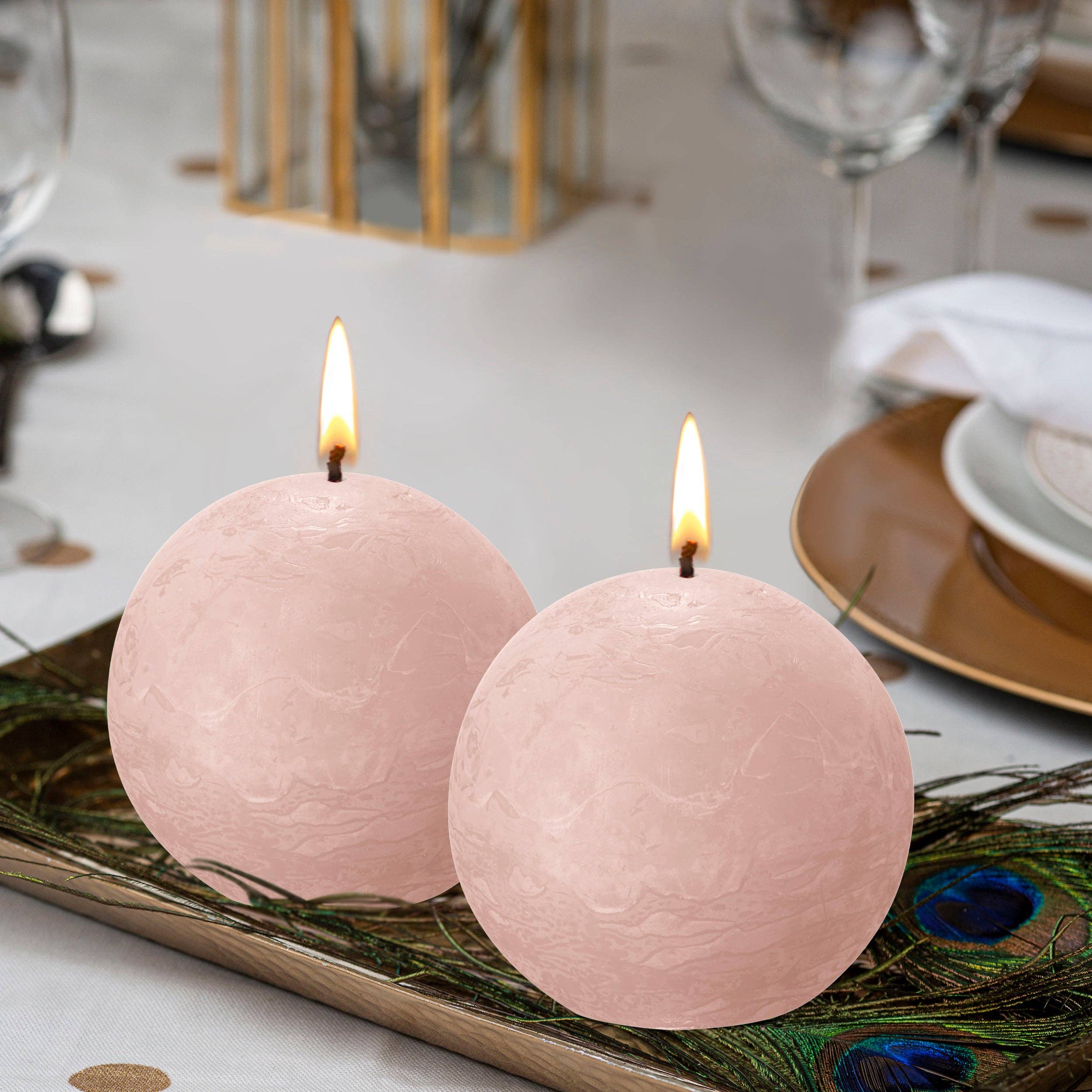 A Misty Pink Rustic 3" Ball Candle made from plant-based wax is lit and displayed on a tray with peacock feathers, set against a backdrop of a dining table laid with plates, glasses, and cutlery.