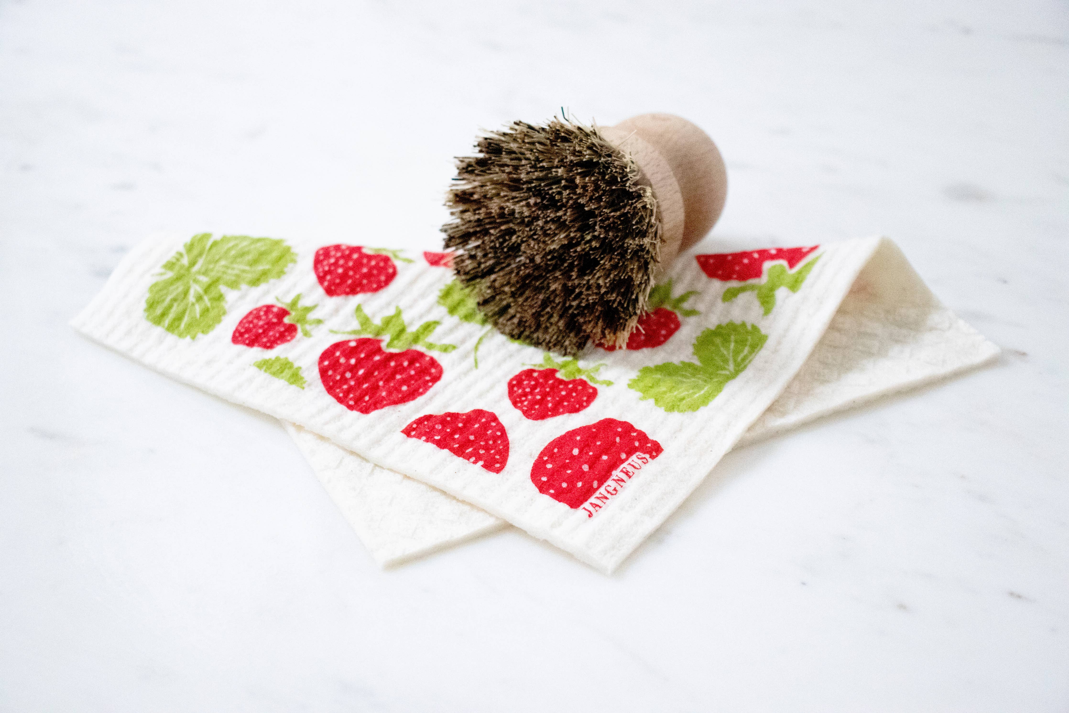 A round wooden scrub brush sits on the Strawberries Swedish Dishcloth, which features red strawberries and green leaves, adding Scandinavian kitchen charm to the white surface.