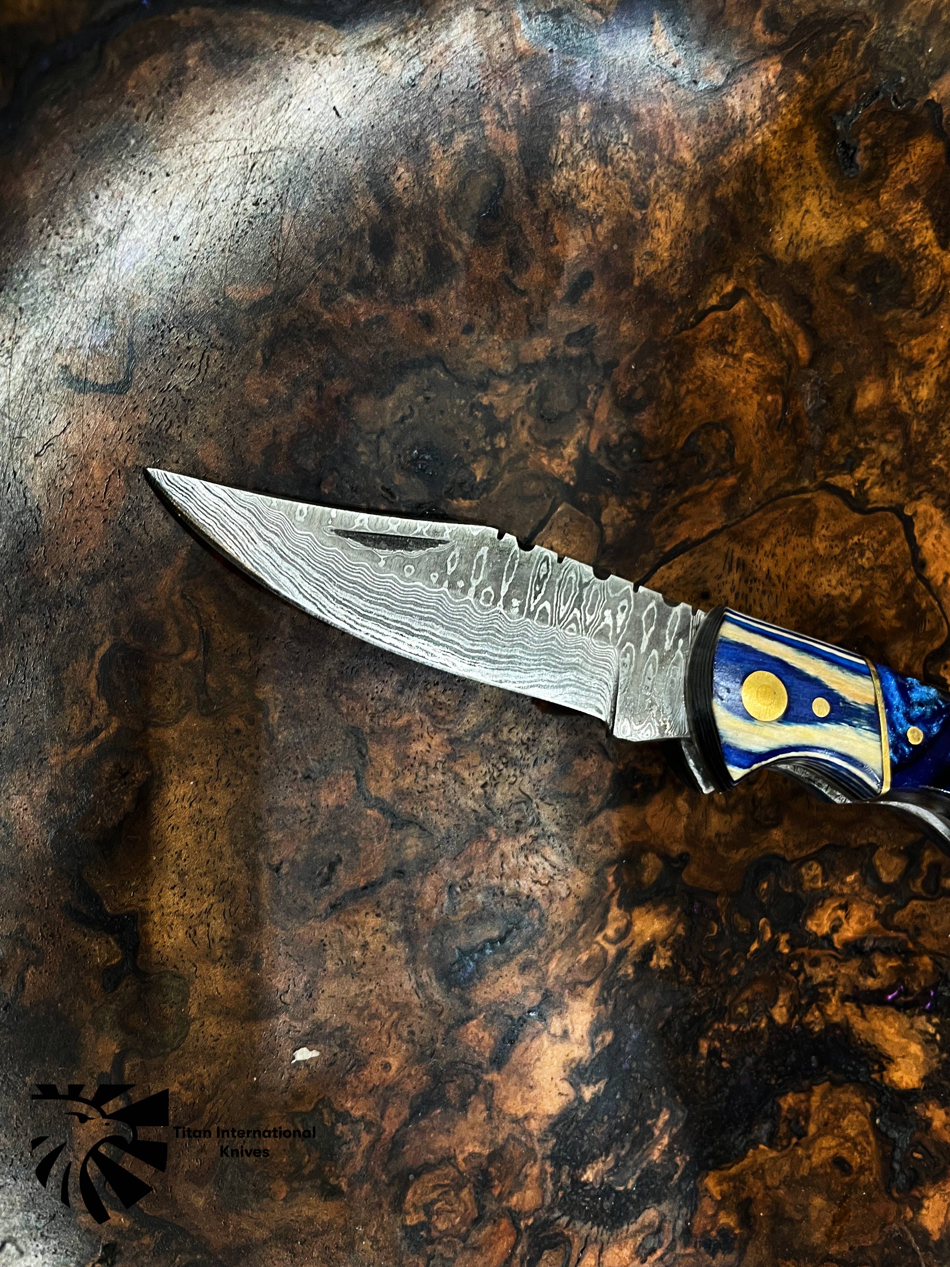 The Titan Celestial Damascus Pocket Knife, featuring a patterned hand-forged blade and a blue-gold handle, rests on a polished dark wood surface.
