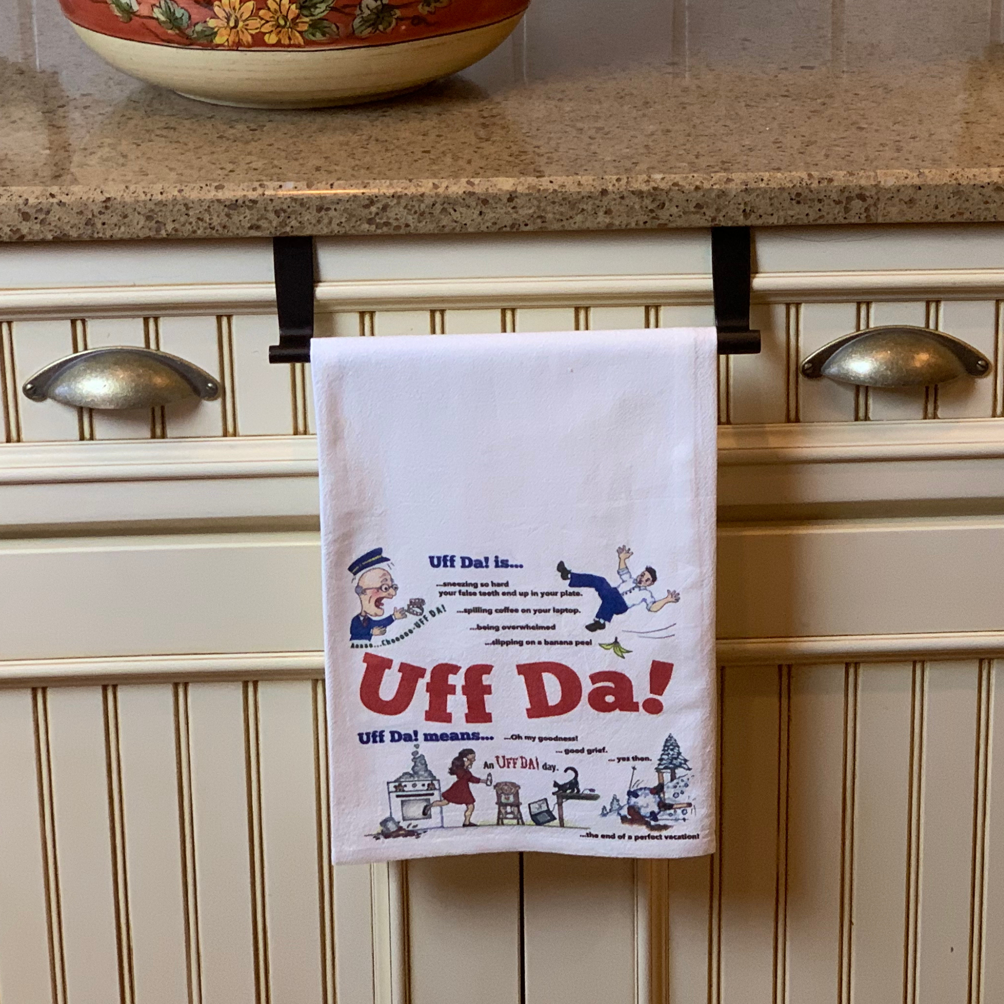 The Tea Towel: UFF Da Norwegian - Swedish Themed Decorative Print Towel hangs on a kitchen cabinet handle, featuring colorful illustrations, humorous definitions, and the phrase "Uff Da!"—a fun Scandinavian kitchen gift.