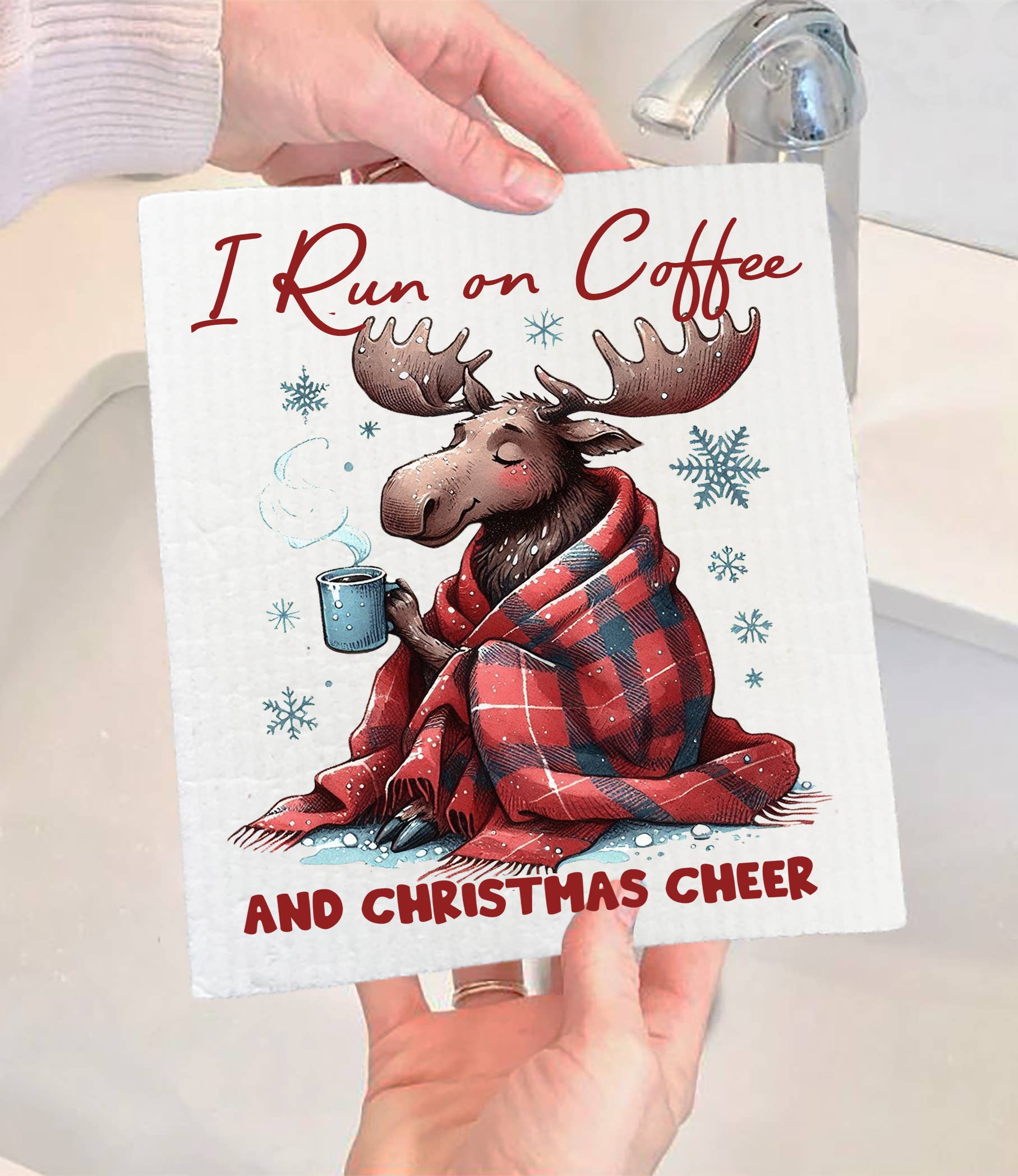 A person holds the Dish Cloth: Christmas Moose I Run on Coffee & Christmas Cheer Swedish Dishcloth, featuring a moose in a plaid blanket with a coffee mug and festive text.