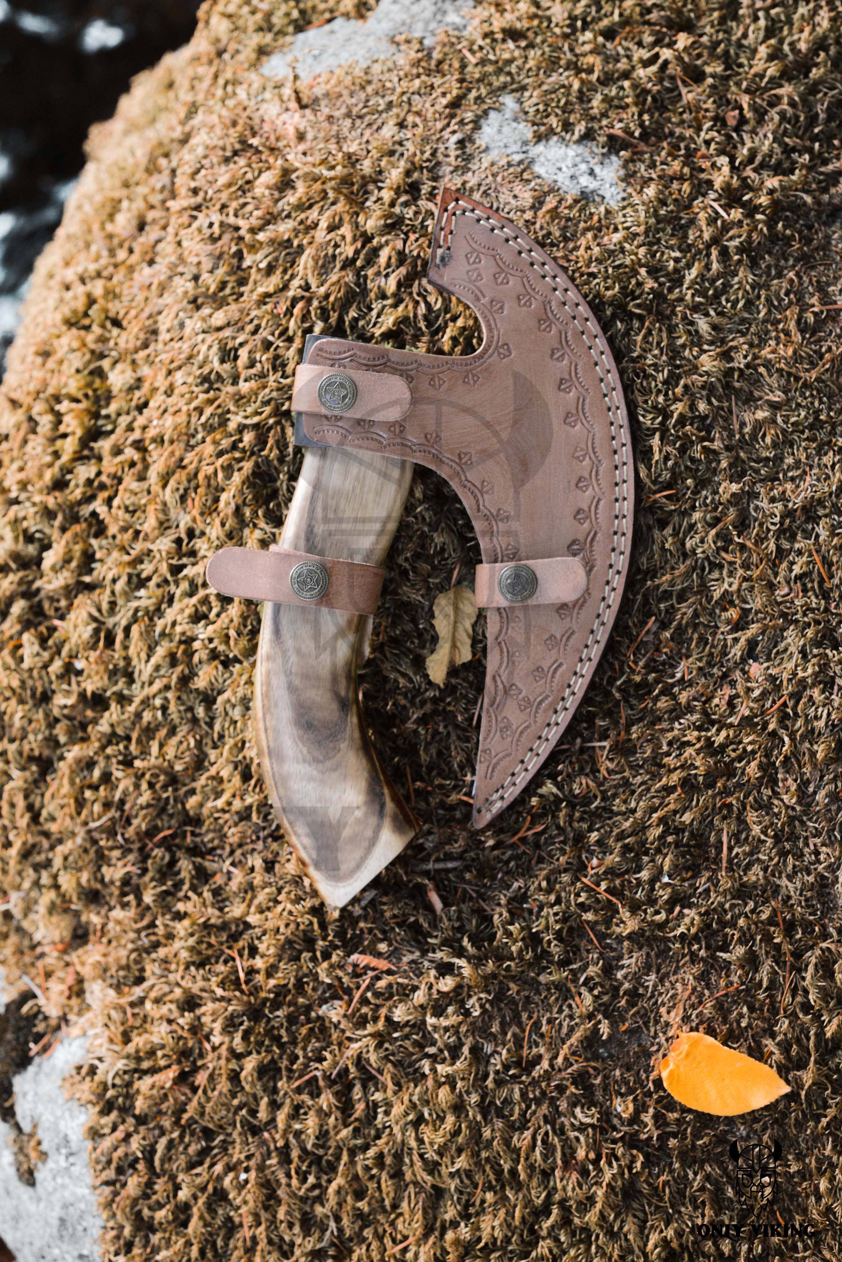 A decorative wooden-handled pizza cutter, featuring a stainless steel blade and engraved leather axe sheath, rests on a moss-covered surface with a small yellow leaf nearby.