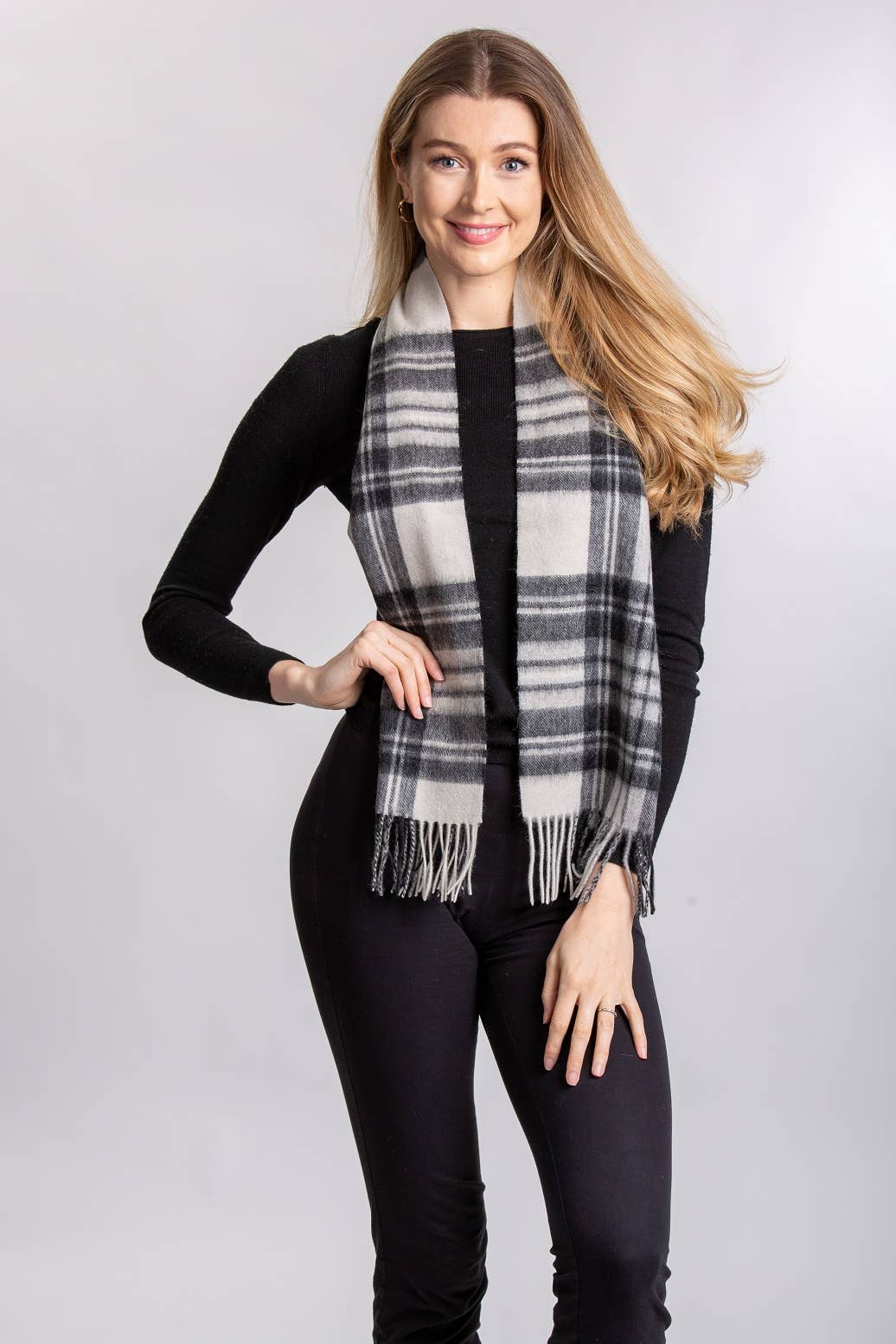 A woman stands and smiles against a plain light background, wearing a black long-sleeve top, black pants, and the Patrick King Stewart Grey Dress Lambswool Scarf.