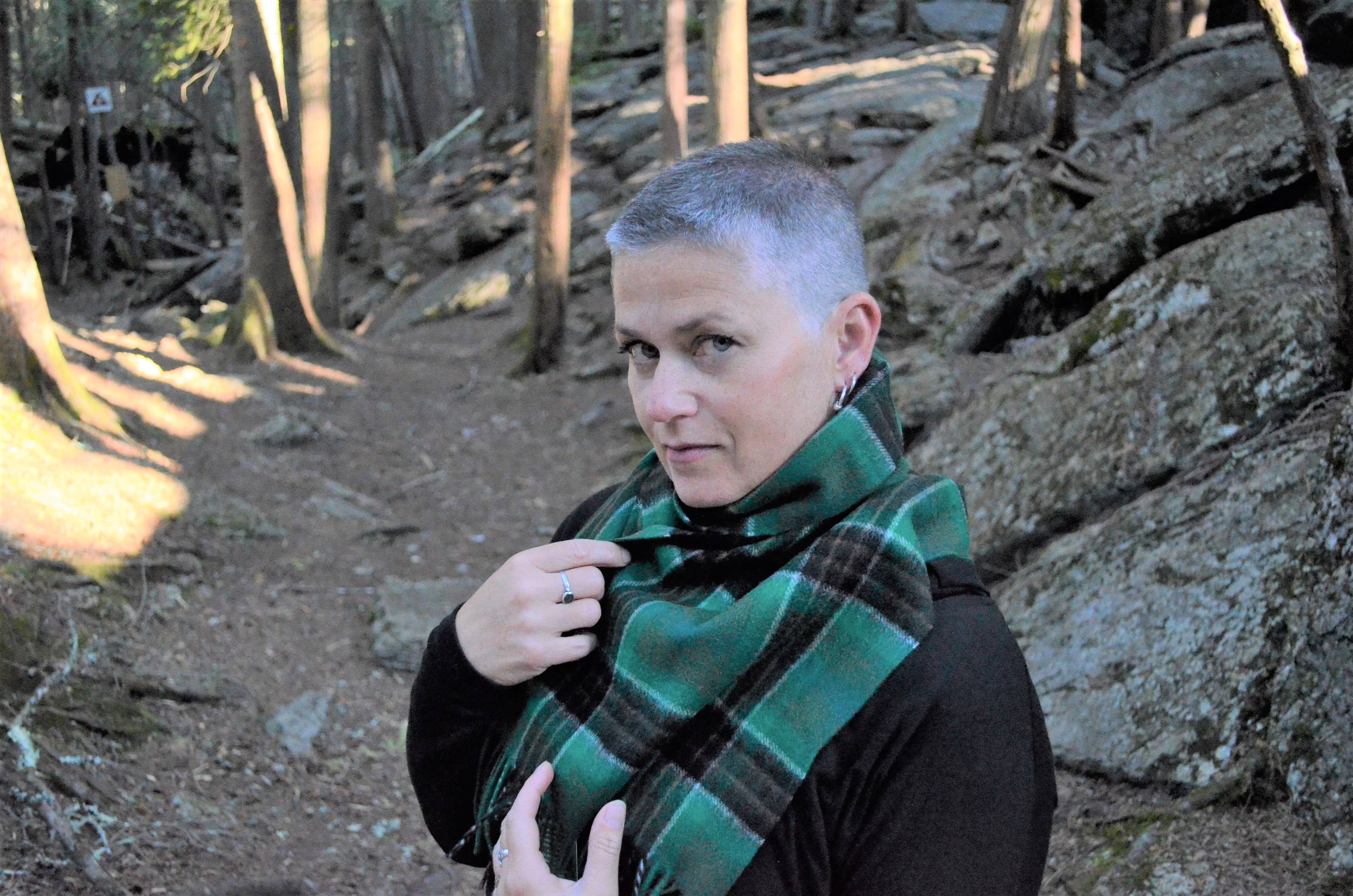 A person with short hair wearing the Spirit of Ireland Lambswool Scarf stands on a rocky forest path, looking directly at the camera.