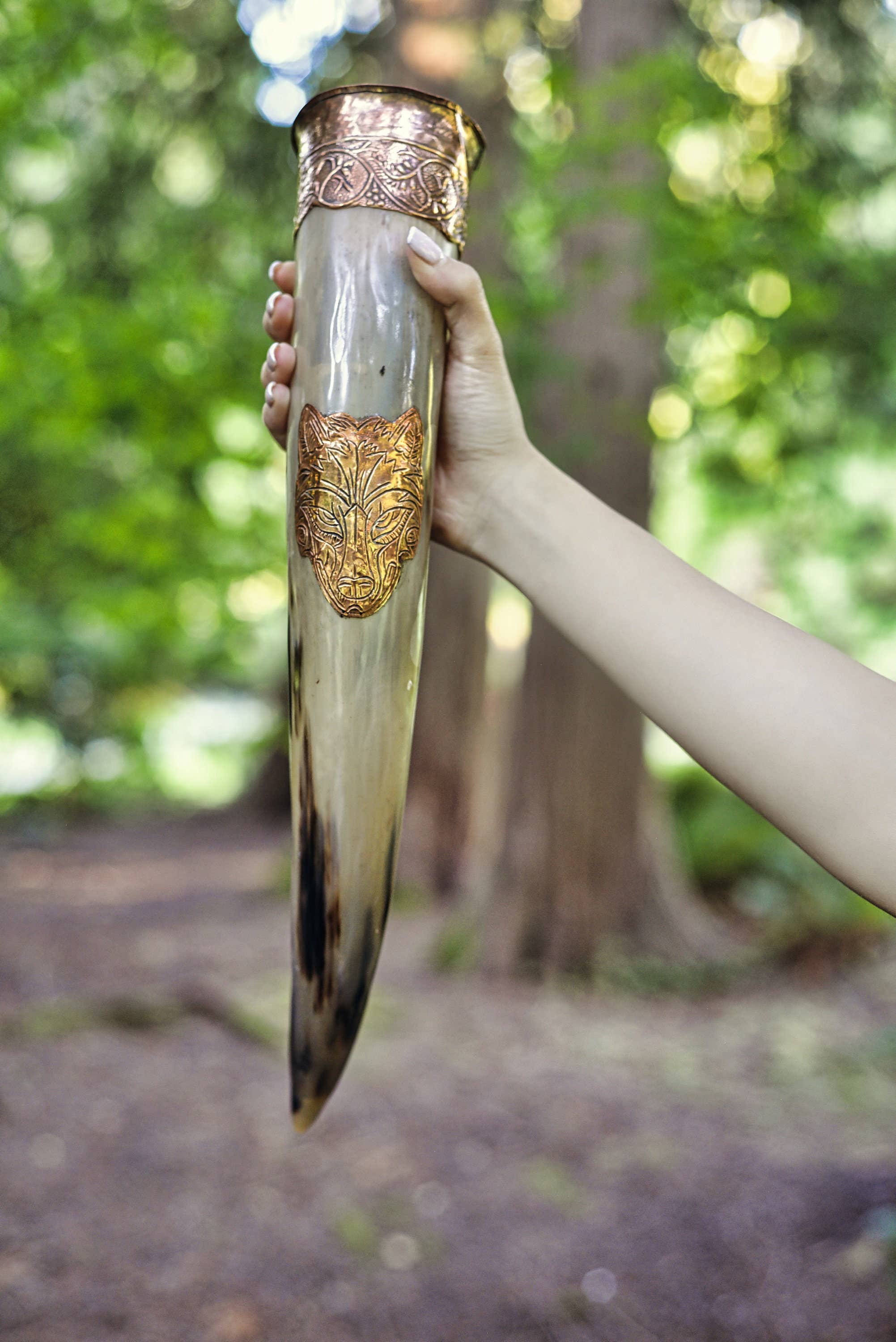 A hand displays the Drinking Horn: Authentic Viking Wedding Gift—an ornate ox horn with gold-colored metal accents and engraved designs, set before a blurred outdoor forest background.