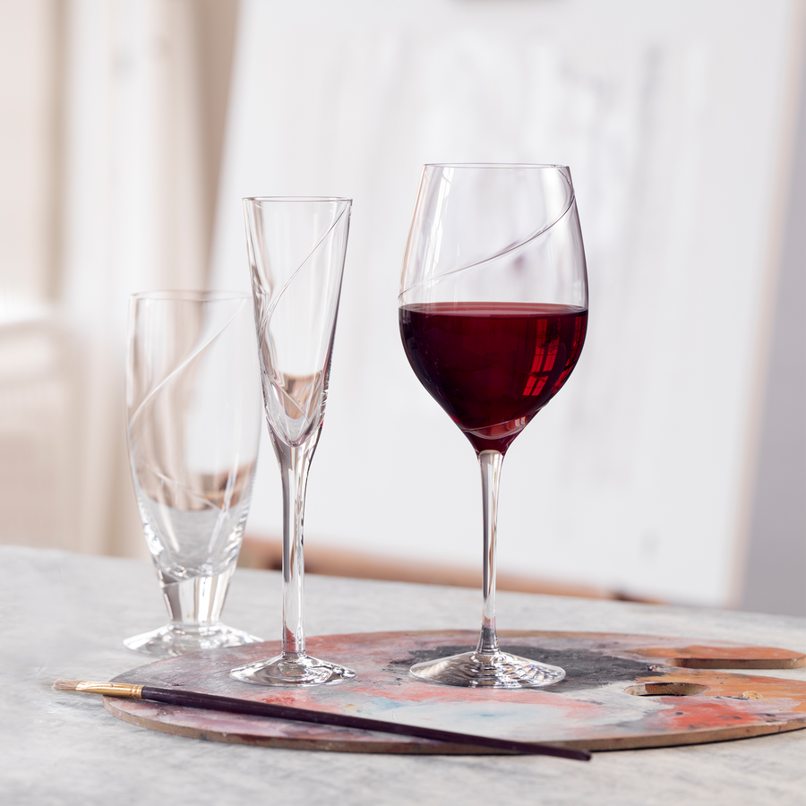 Three glasses—a mouth-blown tumbler, a KostaBoda: Line Flute Champagne Glass, and a wine glass with red wine—are artfully arranged on a painter’s palette next to a paintbrush.