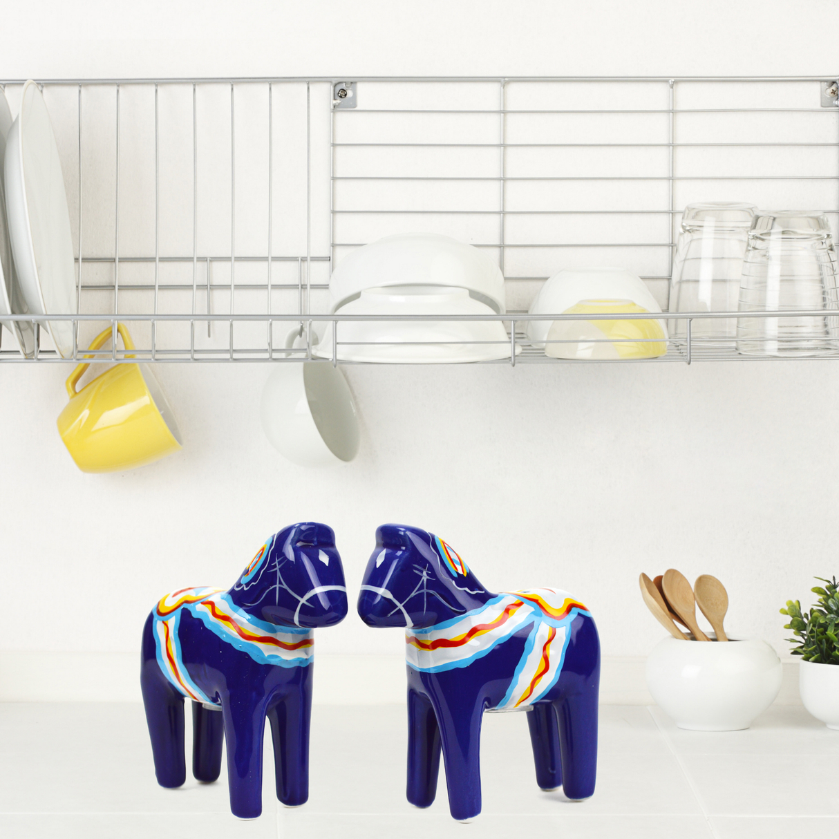 A pair of Blue Swedish Dala Horse Salt and Pepper Shakers bring Scandinavian charm to the white countertop beneath a dish rack filled with plates, bowls, cups, and glasses in this inviting kitchen scene.