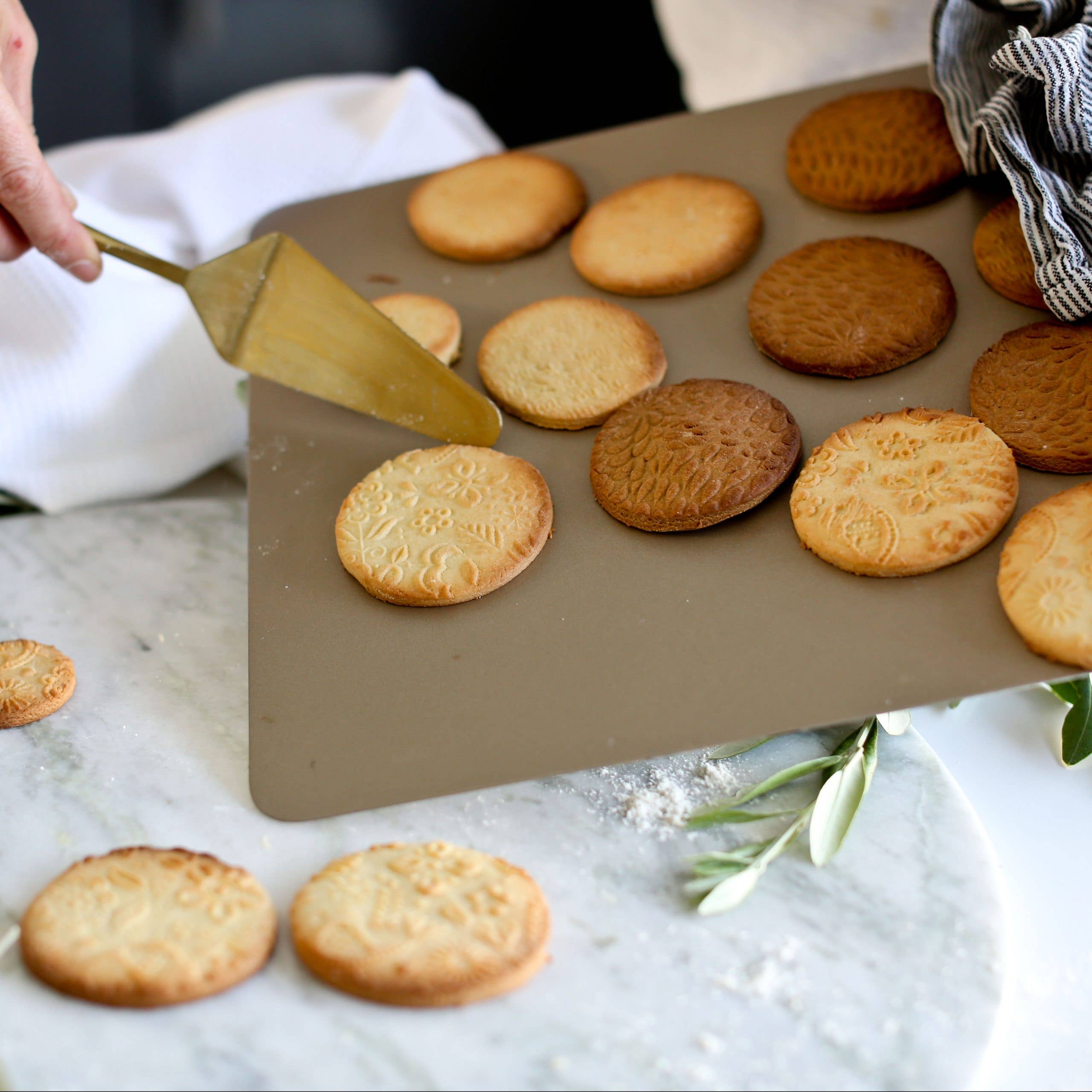 A spatula lifts patterned cookies from a baking tray onto a marble surface. Nearby, the Rolling Pin: Folk Pattern Embossing Rolling Pin Flowers hints at handmade gifts, while a striped cloth and white towel add charm to the scene.