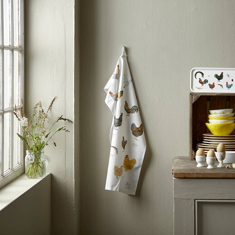 The Tea Towel: Colorful Chickens by Annemarie Haase, made of organic cotton, hangs on a wall next to a window with wildflowers. Nearby, a shelf displays colorful bowls, a tray, and egg cups.