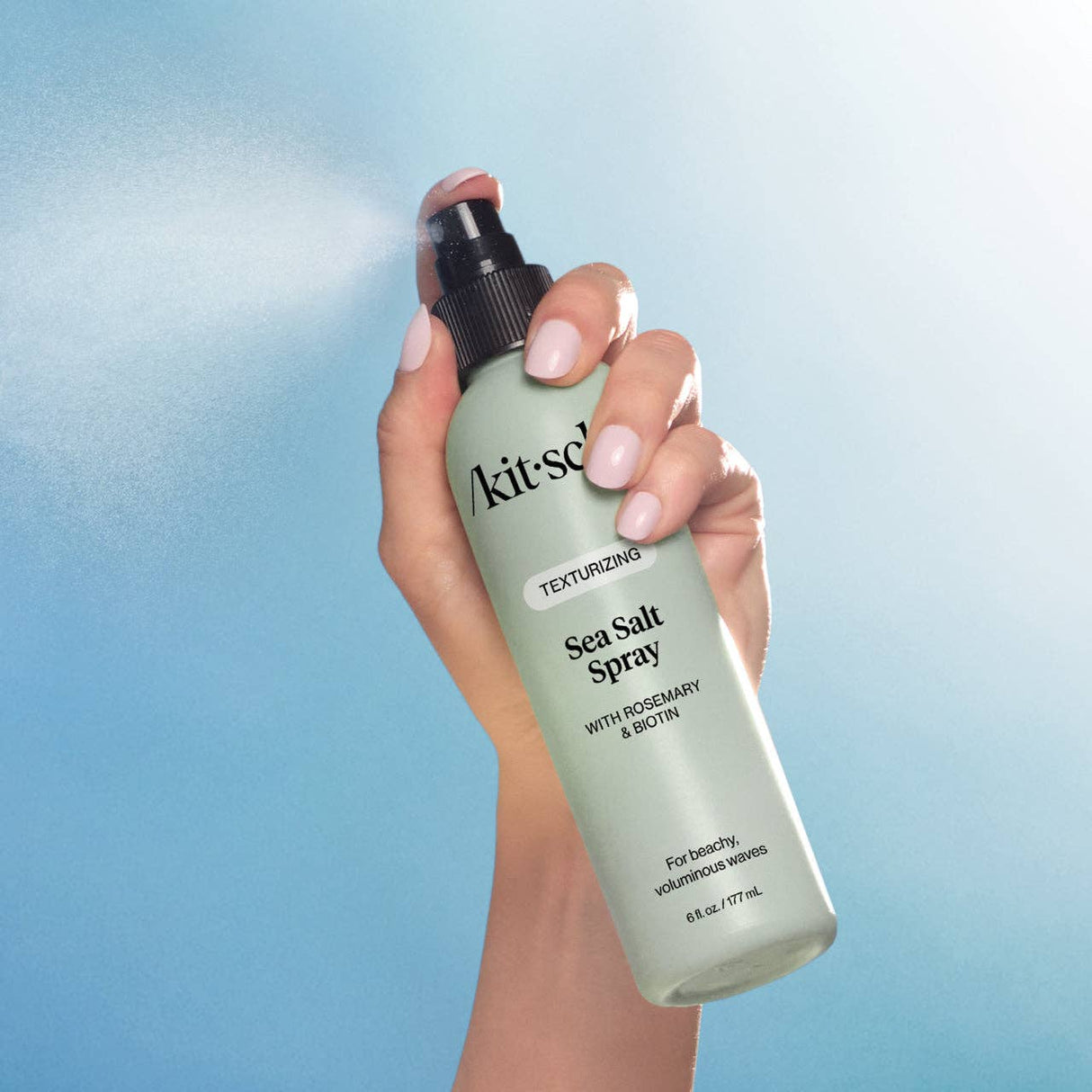 A hand holding and spraying a bottle of KITSCH Spray: Texturizing Sea Salt Hair Spray with Rosemary & Biotin (6 fl oz), a vegan hair product ideal for creating effortless beach waves, against a blue background.
