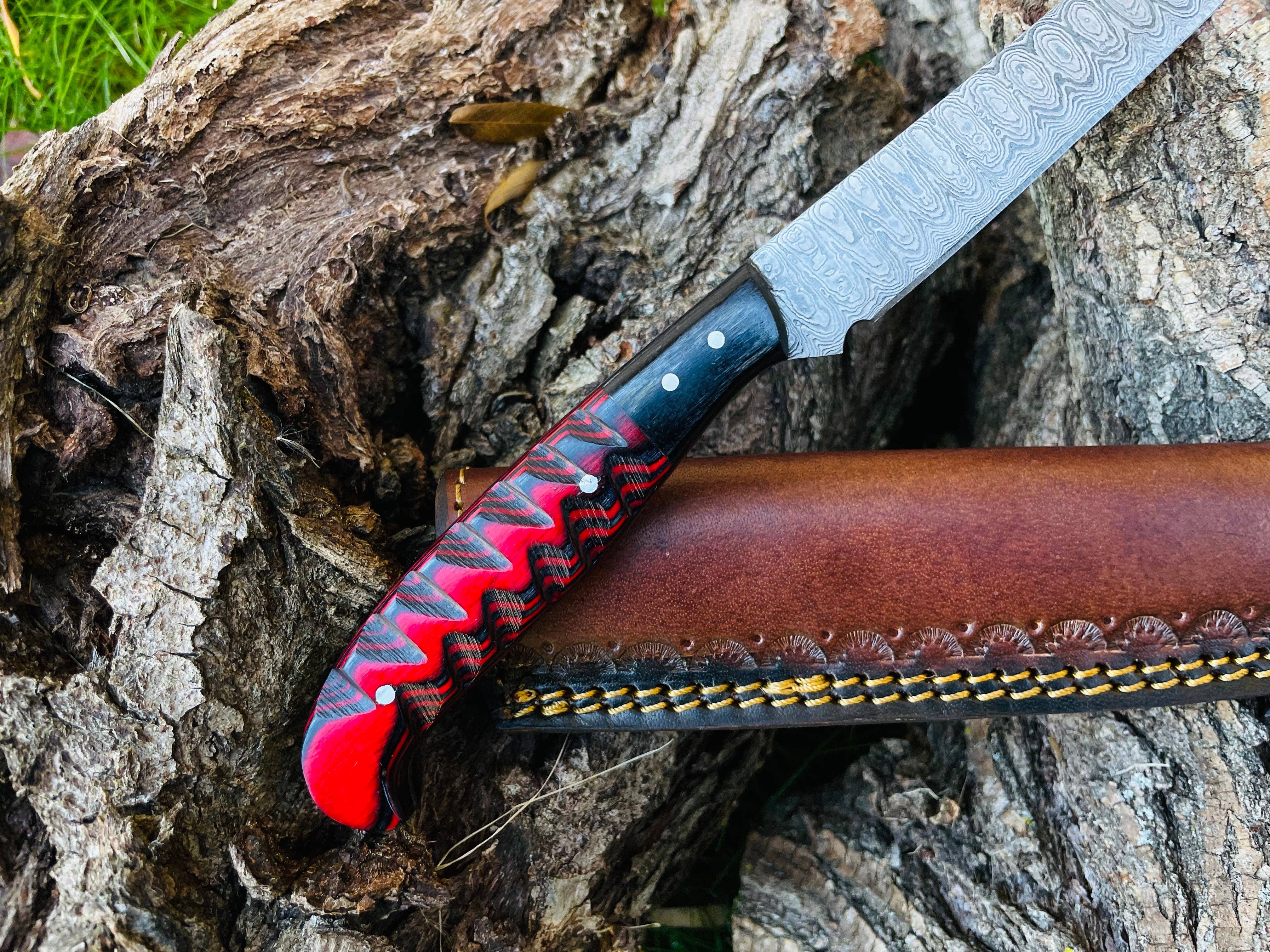 The Knife: Damascus Steel Custom Flay Knife with Craved Grips features a patterned blade and red-black handle, resting on a brown leather sheath atop rough tree bark.